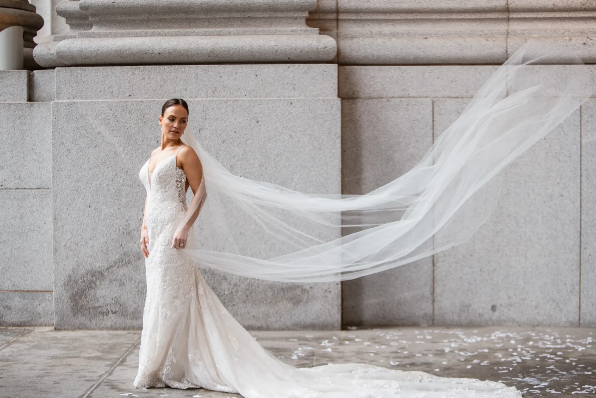 Inhouse Studios Photography, specializing in wedding photography, captures a stunning image of a bride gracefully posing against a grand, stone building. The bride is elegantly dressed in a fitted, lace wedding gown with a long, flowing veil trailing behind her, softly billowing in the breeze. The neutral tones of the stone backdrop enhance the bride's exquisite attire, creating a timeless and sophisticated wedding portrait perfect for Mariage Elegant Canada.