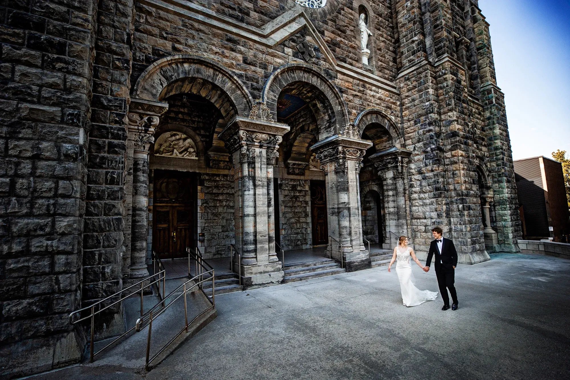 A bride and groom hold hands as they walk in front of a grand stone cathedral with intricate archways and ornate columns, captured by Inhouse Studios Photography. The bride wears an elegant white gown, while the groom is dressed in a sharp black tuxedo. This wedding photography showcases a serene and timeless moment, highlighting the couple's joyous occasion in a historic setting. The image is part of the wedding photography category on Mariage Elegant, Canada, featuring the artistry of Inhouse Studios Photography.