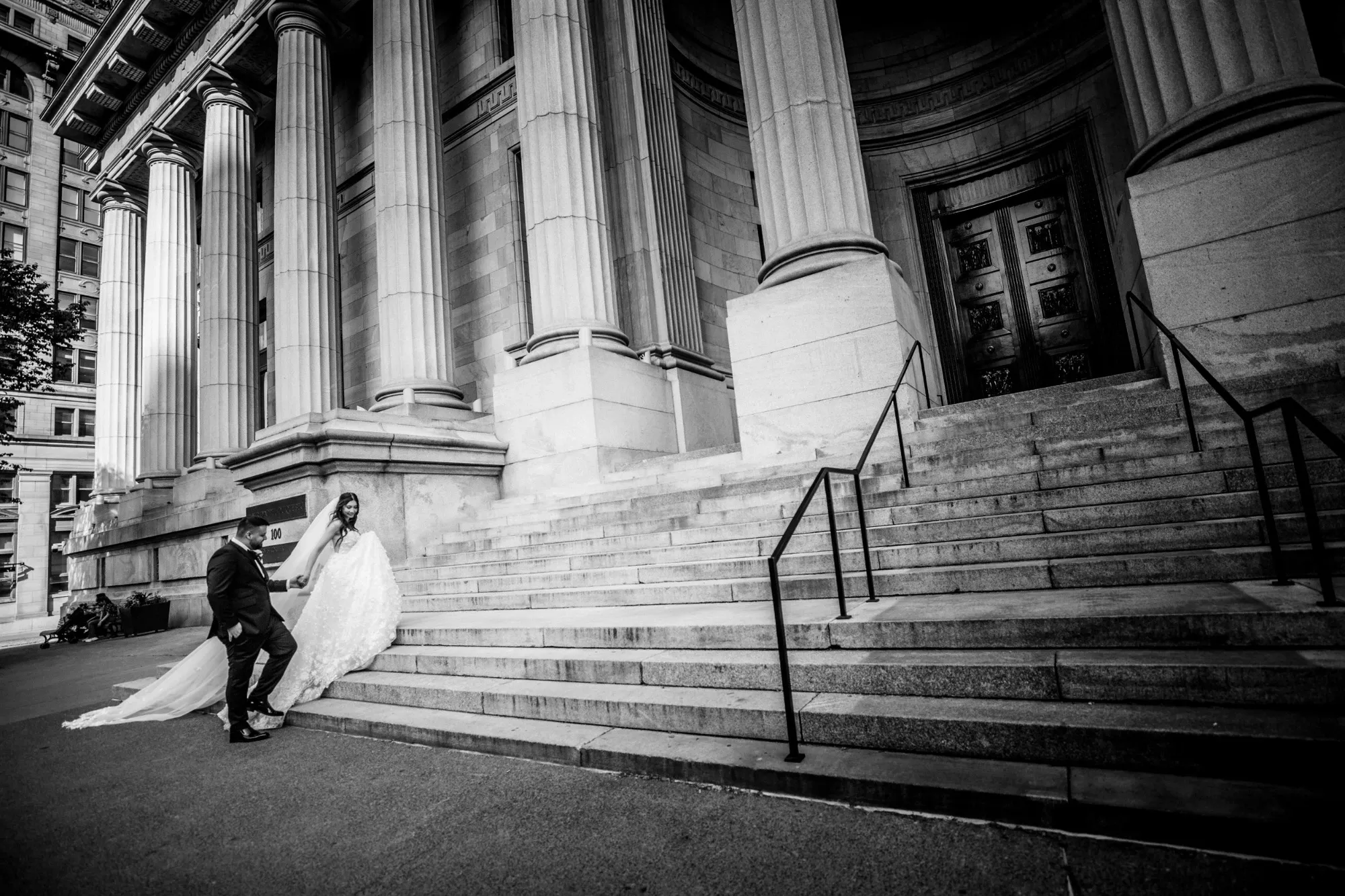 In this black and white image by Inhouse Studios Photography, a newlywed couple is captured in the wedding photography category for Mariage Elegant. The bride, wearing a flowing white gown with a long train, descends the grand stone steps of an impressive, column-lined building. Her groom, in a dark suit, gently holds her hand. The architectural detail of the venue provides a classic and timeless backdrop, enhancing the elegance of the moment. The scene conveys a sense of romance and celebration, ideal for a wedding photography portfolio.