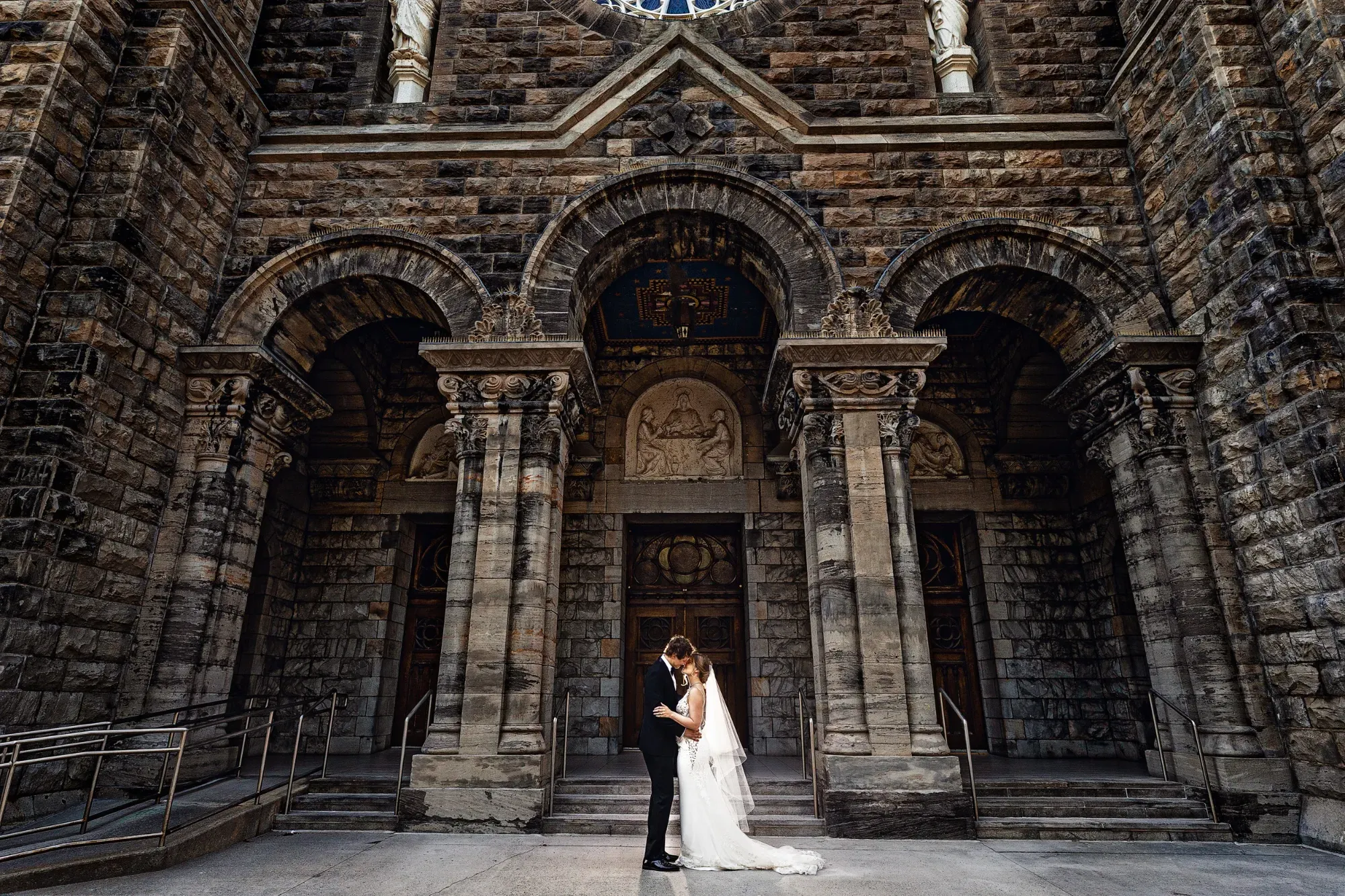 A bride and groom stand intimately in front of a grand, historic stone church facade, showcasing intricate architecture with tall arches and detailed carvings. The bride wears an elegant white gown with a flowing train, while the groom is dressed in a classic black suit. They are positioned in the center, holding hands and gazing at each other, capturing a moment of affection. The sunlight casts gentle shadows, highlighting the textures of the church's stonework. This image, taken by Inhouse Studios Photography, beautifully encapsulates the essence of wedding photography, emphasizing romance and architectural elegance.