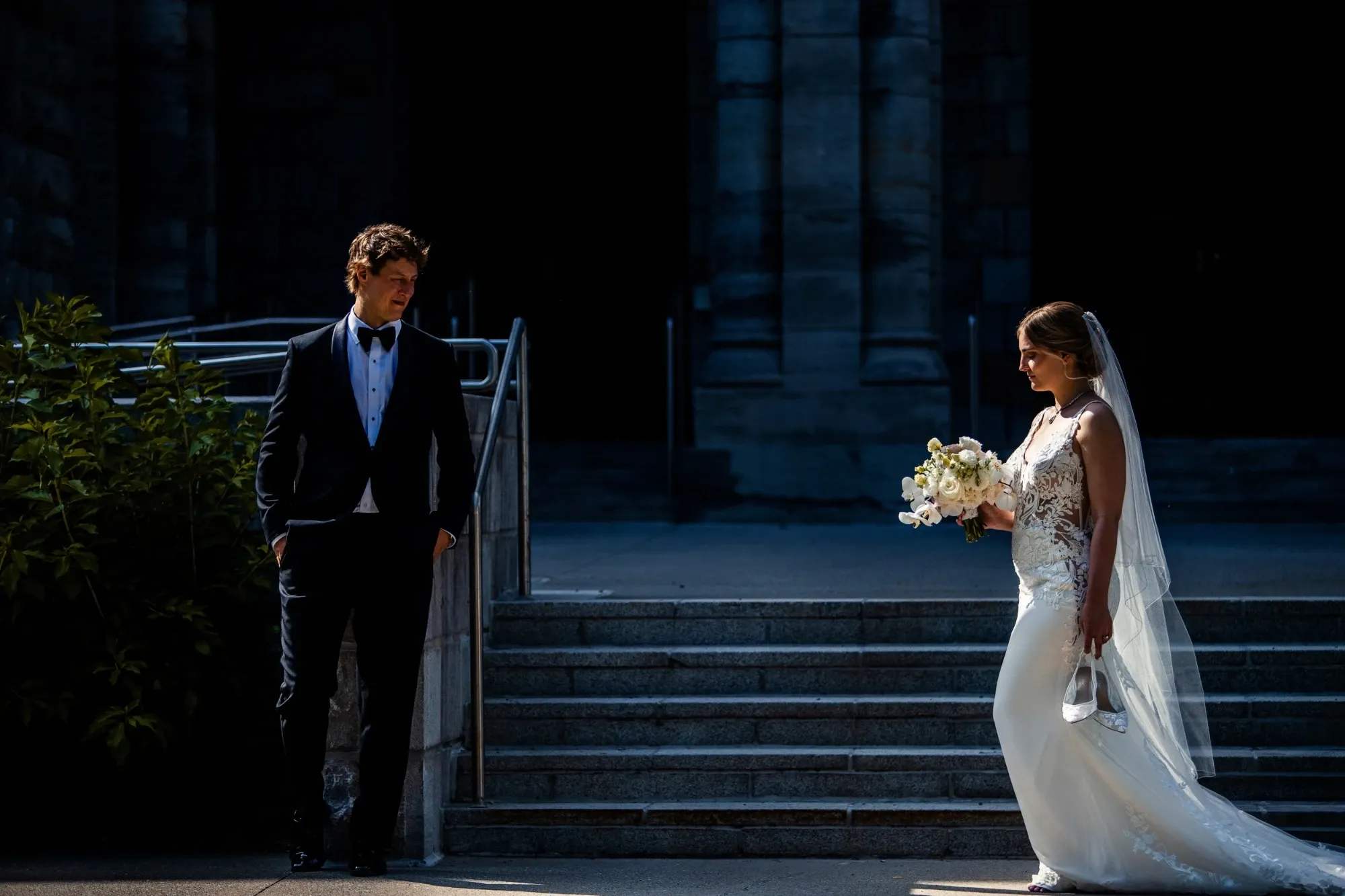 The image shows a bride and groom in an elegant, dimly lit outdoor setting. The bride is wearing a beautiful white lace wedding gown and holding a bouquet of white flowers, while the groom stands opposite her in a classic black tuxedo with a white shirt and black bow tie. They are positioned in front of a grand stone staircase, which adds a touch of sophistication to the scene. This image captures a romantic moment, perfect for showcasing the skills of Inhouse Studios Photography, a vendor specializing in wedding photography within the Mariage Elegant listings in Canada.