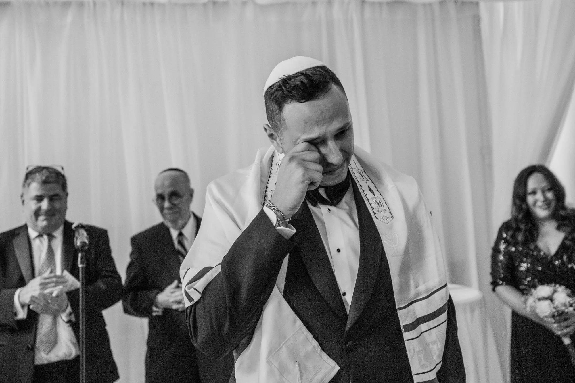 Inhouse Studios Photography captures an emotional moment in the wedding ceremony, focusing on a groom standing in front of a white draped backdrop. The groom is dressed in a formal black tuxedo with a white kippah and tallit, wiping away tears of joy. In the background, several guests, including two men in suits and a woman in a sequined dress, watch the scene with expressions of happiness. This image, showcasing wedding photography, highlights the heartfelt emotions present during a marriage celebration, perfectly aligning with the services of Inhouse Studios Photography under the wedding photography category on Mariage Elegant, Canada.