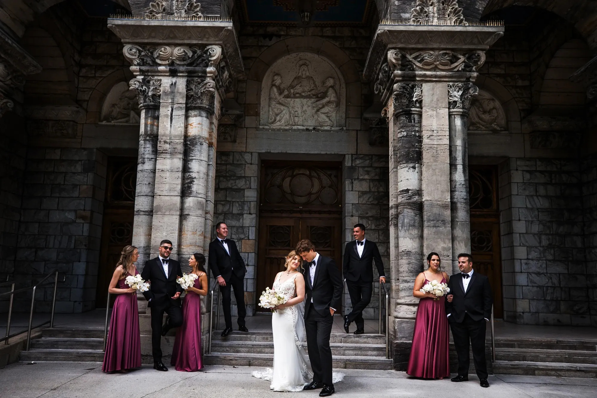 This image from Inhouse Studios Photography, highlighting their expertise in wedding photography, captures a sophisticated wedding party posed outside a grand, historic stone building with ornate columns. The couple stands gracefully at the center, with the bride holding a beautiful bouquet, while the bridal party, dressed in elegant attire with the bridesmaids in matching burgundy dresses, complements the scene. The mood is romantic and timeless, aligning with the elegant wedding theme.