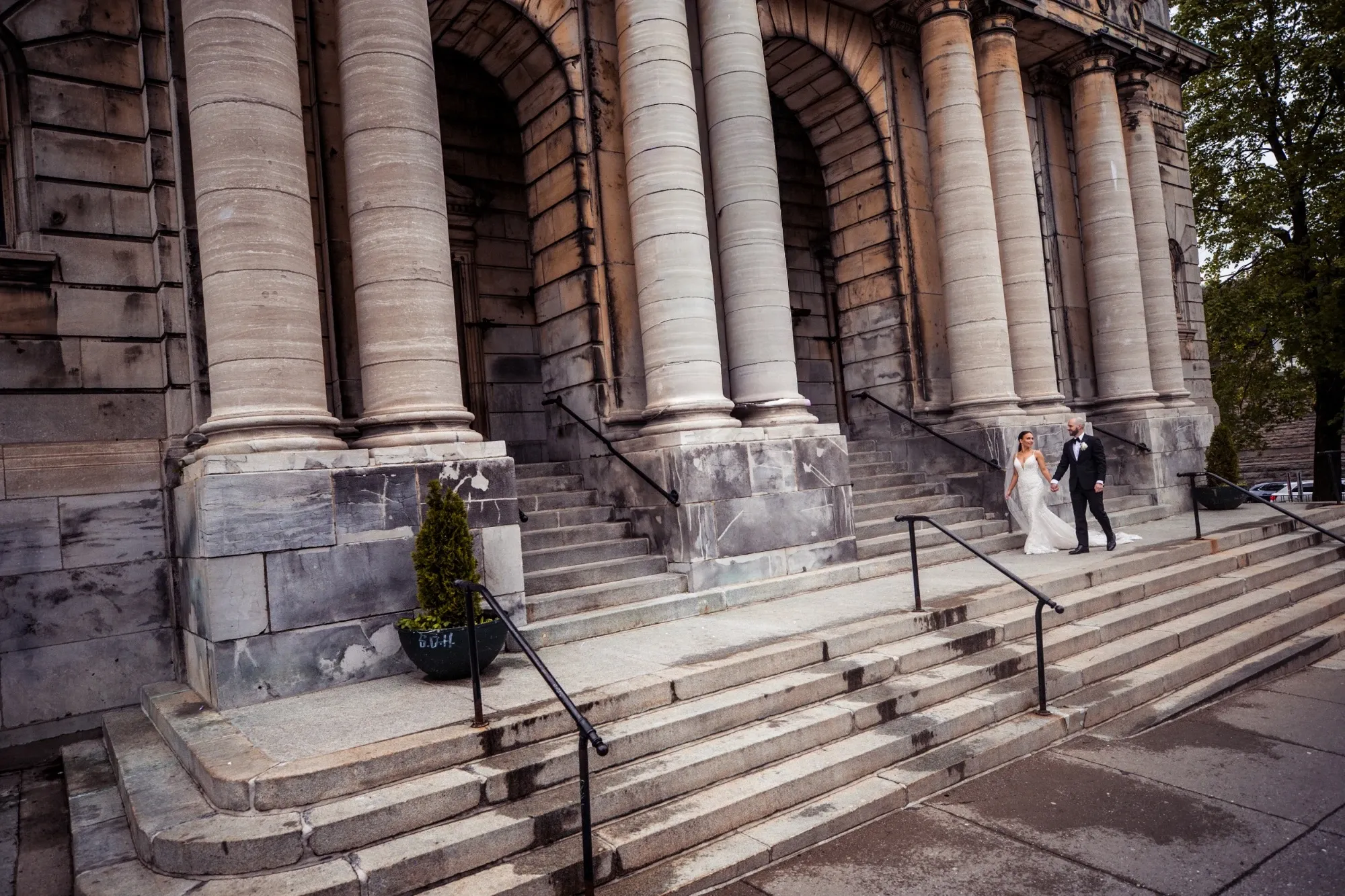 A newlywed couple walks hand-in-hand down the grand stone steps of a historic, columned building, evoking a timeless elegance. The bride, dressed in a traditional white gown, carries a bouquet, while the groom in a classic black suit, complements her grace. This captivating image by Inhouse Studios Photography falls under the Wedding Photography category on Mariage Elegant, Canada, showcasing their ability to capture romantic and sophisticated moments in iconic settings. The architecture's grandeur adds a majestic backdrop to the couple's joyful journey.