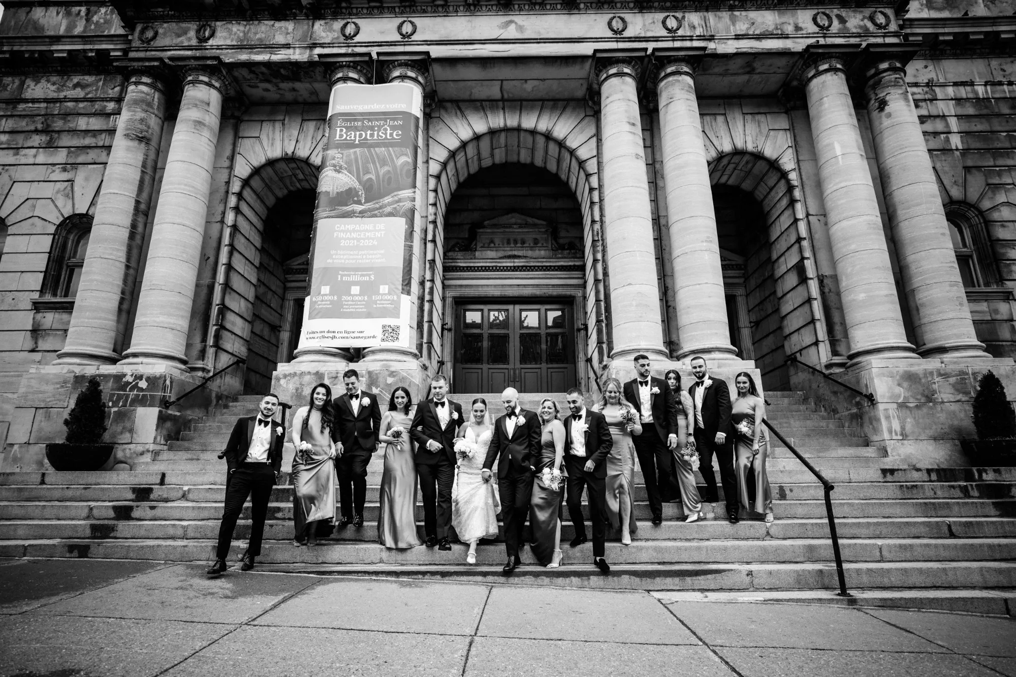 A wedding party poses on the steps of a grand, historic building with large stone columns in the background. The group includes bridesmaids in elegant dresses and groomsmen in formal suits, all smiling and standing close together. This image showcases the work of Inhouse Studios Photography, capturing a joyful moment in wedding photography. The scene conveys a sense of timeless celebration, with the architectural details adding a majestic backdrop to the occasion. The photo is suitable for the vendor listing on Mariage Elegant, Canada.