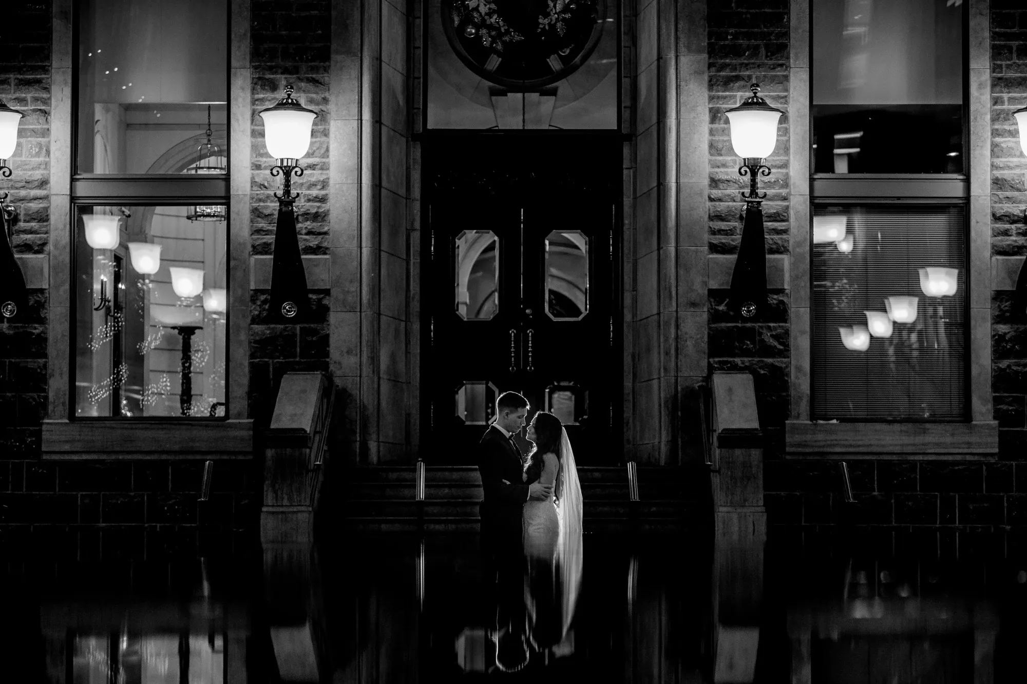 A black and white photograph showcases a bride and groom embracing in front of an elegant building, beautifully captured by Inhouse Studios Photography, a vendor in the Wedding Photography category. The couple stands illuminated under glowing street lamps that frame the entrance, creating a romantic and timeless atmosphere. The reflection of the couple and the ornate architecture in a still pool of water below adds a dramatic and artistic effect, perfect for a memorable wedding moment.