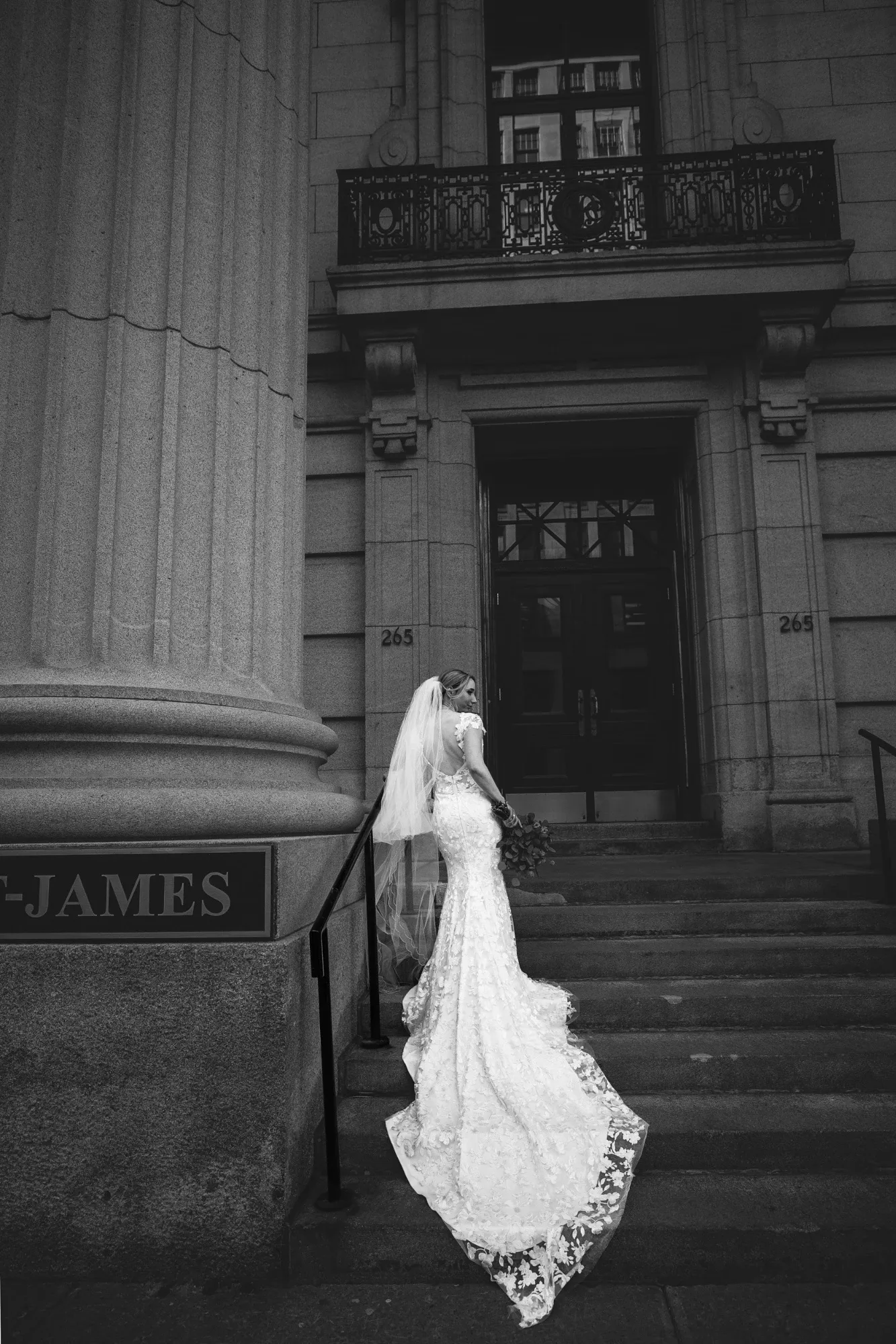 This black and white photograph by Inhouse Studios Photography captures a bride elegantly posed on the steps of a grand building, highlighting the wedding photography category. The bride is wearing a stunning, intricate lace gown with a long train and a veil, creating a timeless and sophisticated look. The setting features tall columns and architectural details, adding to the grandeur of the scene. The image reflects the elegance and beauty that Mariage Elegant, Canada's wedding planning service, aims to promote through its vendor listings.