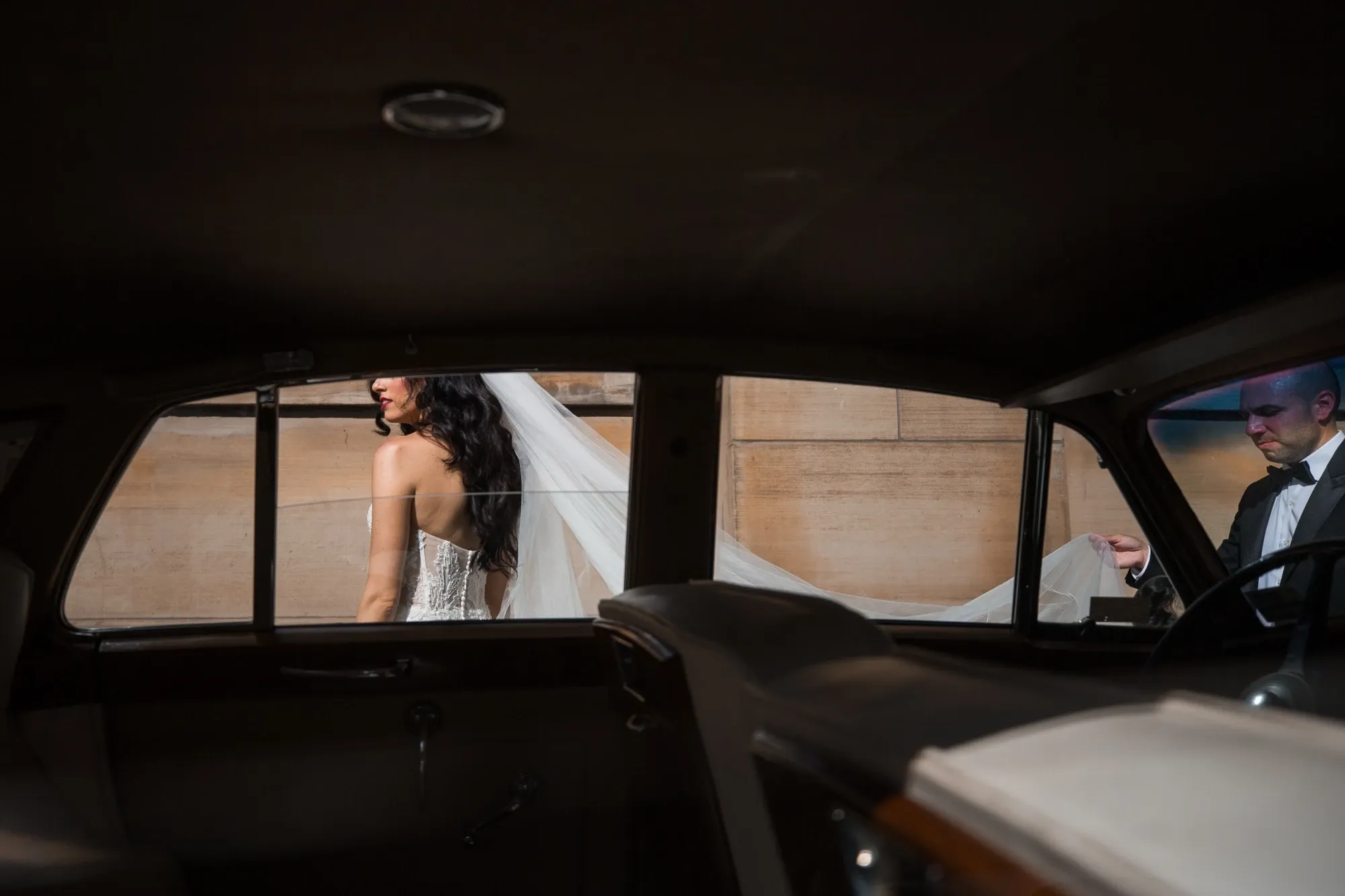 A bride in a white, strapless lace wedding gown is captured walking alongside a vintage car, her long veil being elegantly carried by a person in a tuxedo. This striking scene, photographed by Inhouse Studios Photography, exemplifies their expertise in capturing intimate and stylish moments in the wedding photography category. The image highlights the elegance and sophistication expected in a wedding setting, showcased against a classic backdrop.
