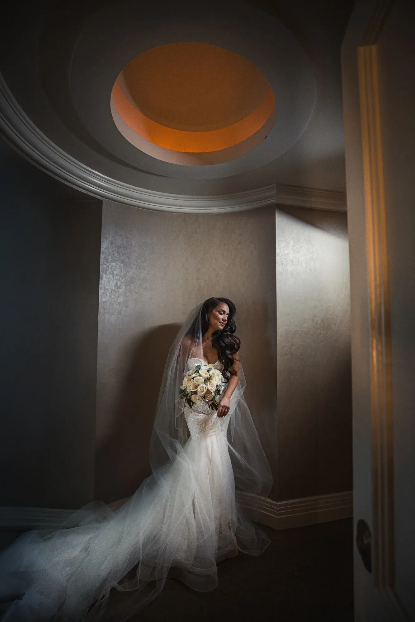 The image features a bride elegantly standing under a soft spotlight in a beautifully curved alcove with a decorative round ceiling, captured by Inhouse Studios Photography. She is wearing a flowing white wedding gown with a long, delicate train and holding a bouquet of pale roses, highlighting the category of wedding photography. Her veil drapes gracefully over her shoulders, enhancing the romantic atmosphere. The setting exudes an intimate and serene ambiance, perfectly suited for Mariage Elegant's wedding planning and vendor listings in Canada.