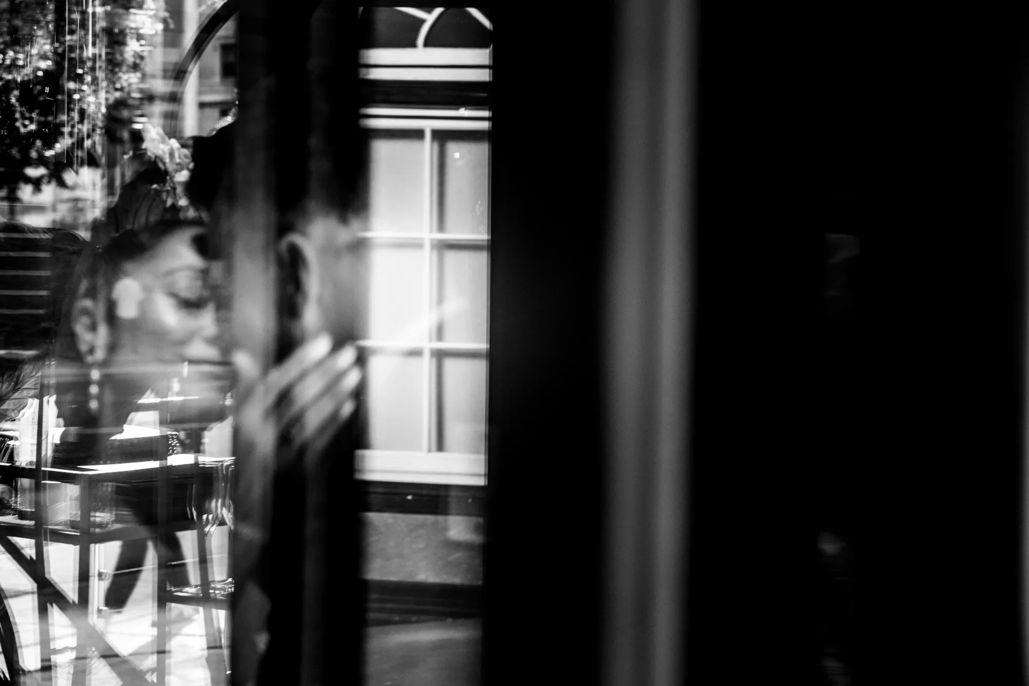 This black-and-white image by Inhouse Studios Photography captures an intimate and emotional moment in the wedding context. The photograph shows a bride, slightly obscured and reflected in a windowpane, creating an artistic and dreamy effect. Her expression suggests deep contemplation, complemented by the soft light and shadow play across her face. The background features a window with geometric patterns, adding a touch of elegance and sophistication to the composition. This image wonderfully encapsulates the essence of wedding photography, showcasing both artistic style and emotional depth.