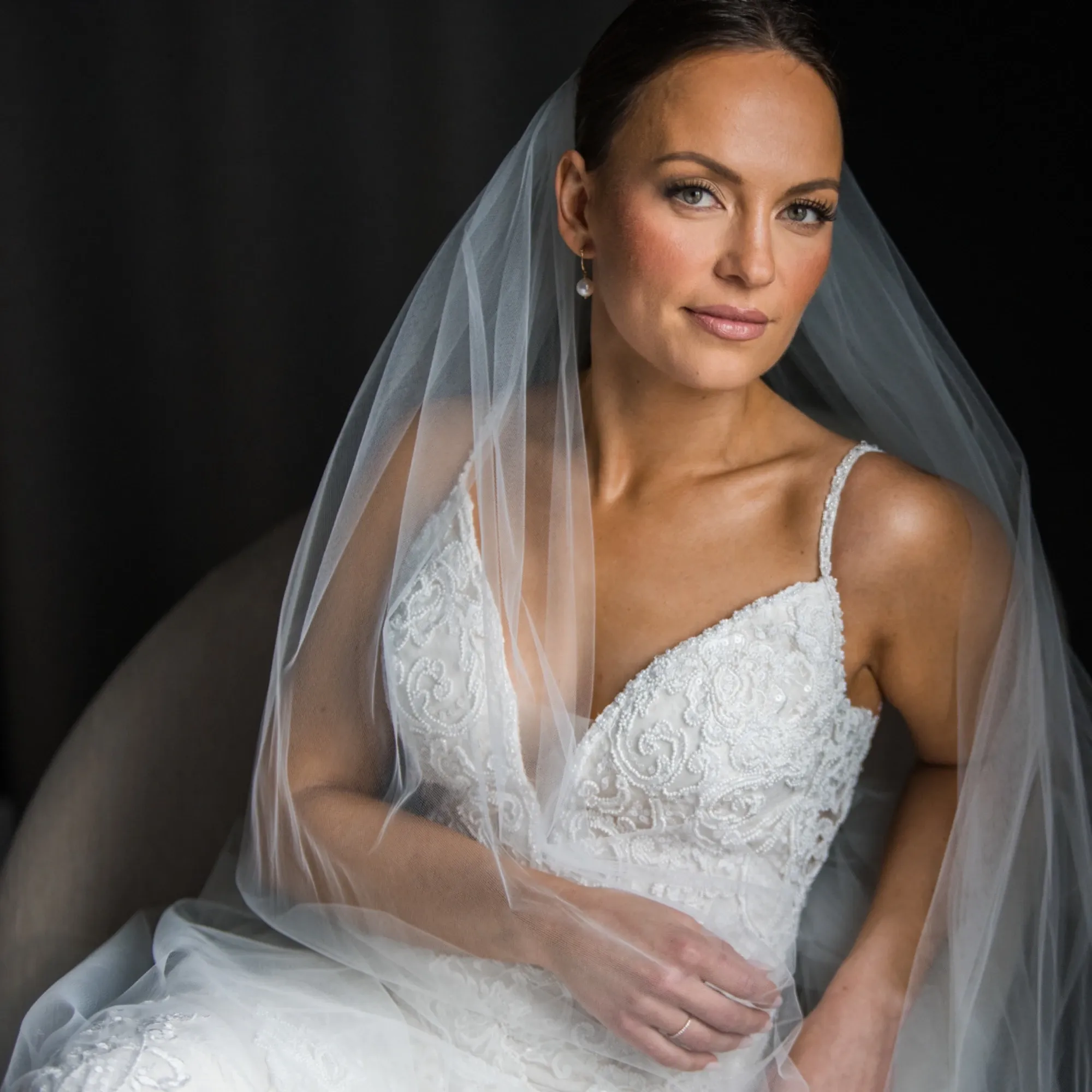 The image features a bride in an elegant, intricately detailed white wedding gown, sitting gracefully and looking confidently into the camera. Her veil delicately drapes over her shoulders, enhancing the sophistication of the scene. The lighting highlights her poised expression and the fine details of her lace dress. This photo, expertly captured by Inhouse Studios Photography, exemplifies their skill in wedding photography, focusing on creating timeless and elegant bridal portraits.
