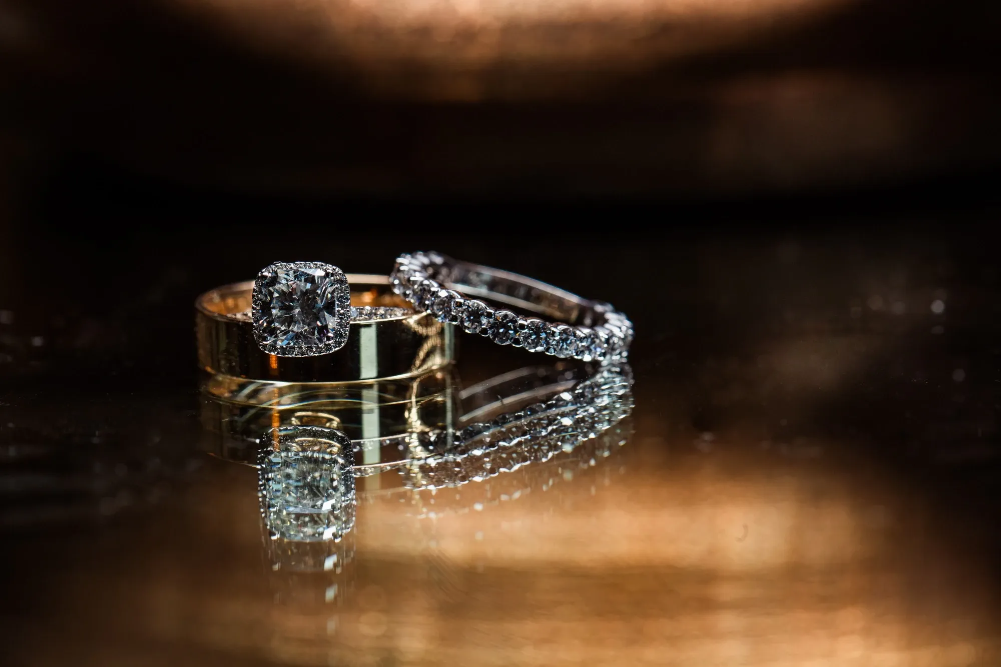 The image showcases two beautifully crafted wedding rings placed elegantly on a reflective surface. One ring features a stunning diamond engagement ring with a halo setting, while the other is a classic gold band. The background is softly blurred, creating a warm, romantic ambiance. This captivating photograph is captured by Inhouse Studios Photography, a vendor specializing in wedding photography, and is perfect for conveying elegance and commitment for Mariage Elegant, Canada.