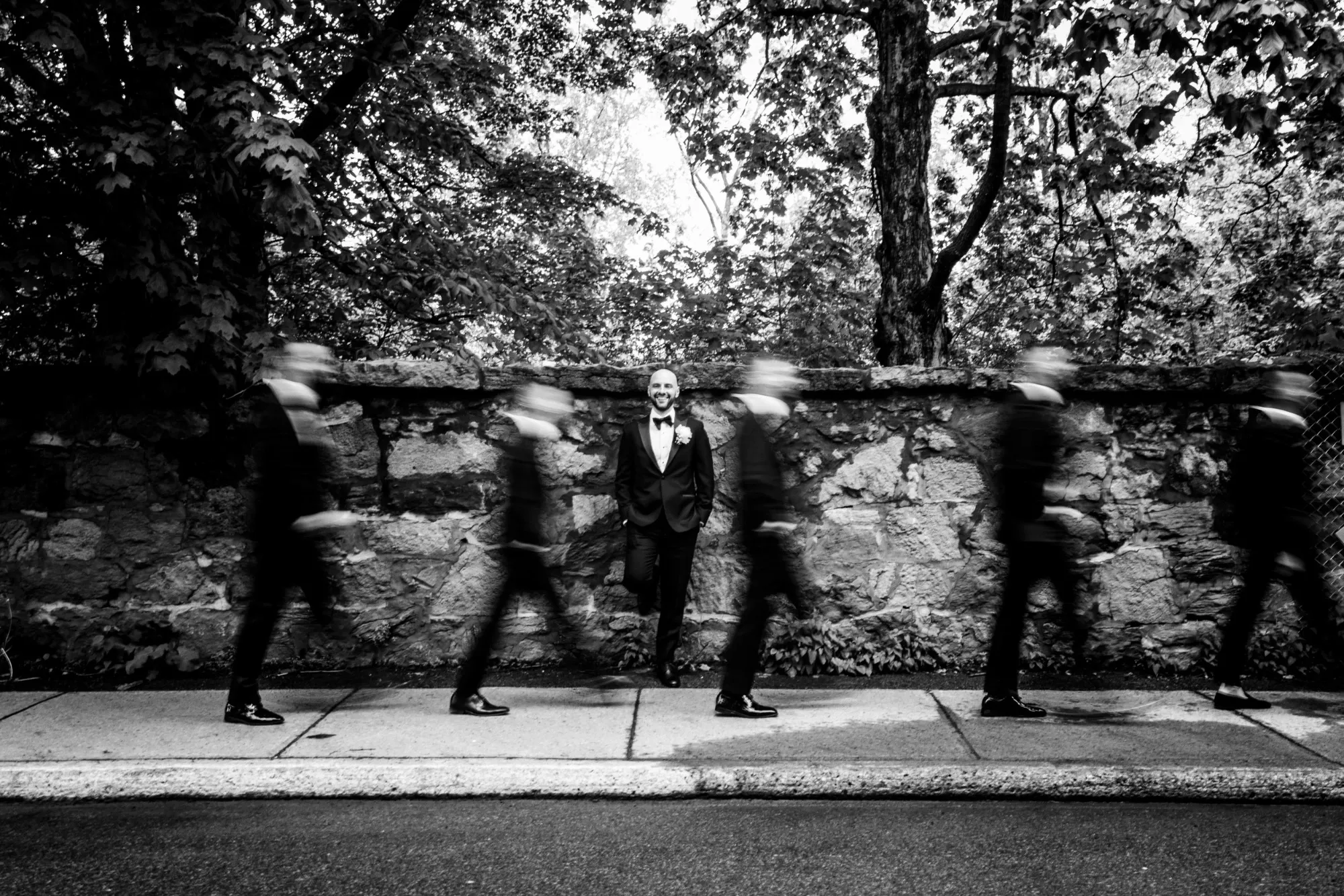 The image features a groom in formal attire, standing confidently against a rustic stone wall, surrounded by a lush, tree-filled background. He is captured in a serene pose while groomsmen, dressed in similar suits, briskly walk in front of him, creating a striking motion blur effect. This black-and-white photograph, presented by Inhouse Studios Photography, showcases their expertise in capturing unique and dynamic wedding moments. The photo highlights the artistry of wedding photography, emphasizing both the stillness and motion of a wedding day, suitable for Mariage Elegant in Canada.