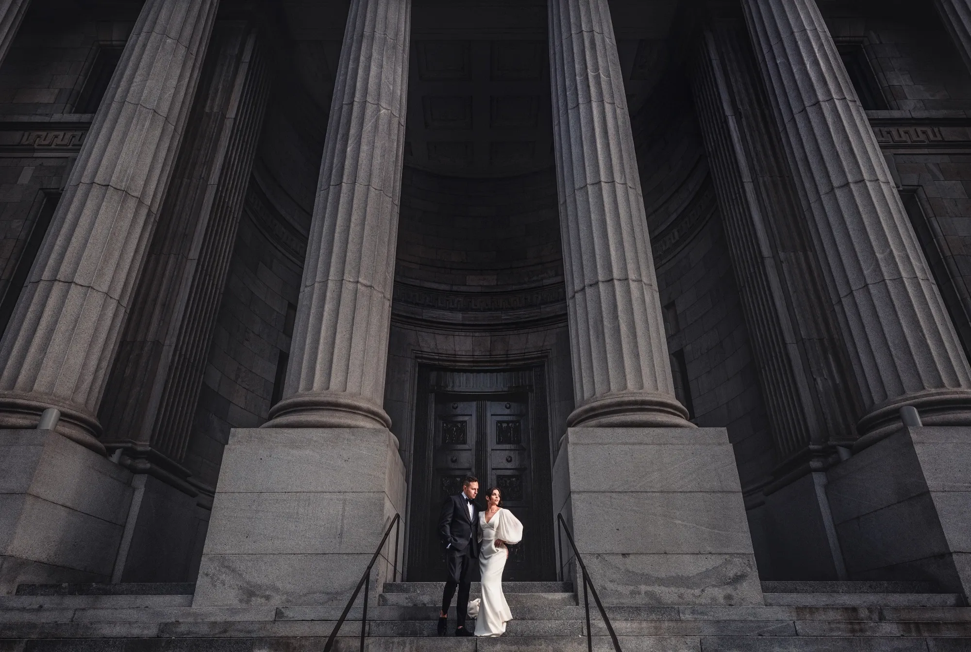 A newlywed couple poses on the steps of a grand building with towering stone columns, showcasing Inhouse Studios Photography's expertise in wedding photography. The bride is elegantly dressed in a white gown, while the groom is in a dark suit. The image captures the dramatic architecture and classic romance, perfect for a wedding setting. This photo exemplifies the high-quality offerings of Inhouse Studios Photography, listed on Mariage Elegant, Canada.