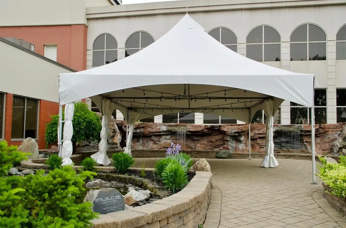 La salle de réception Plaza Rive Sud présente un élégant pavillon blanc en extérieur, parfait pour les mariages. Le pavillon est érigé sur une terrasse pavée entourée de végétation luxuriante, incluant des plantes vertes et des fleurs violettes. En arrière-plan, un mur décoratif en pierre avec des cascades ajoute une touche de sophistication et de sérénité. Cette scène naturelle et bien aménagée offre un cadre idyllique pour les célébrations de mariage.