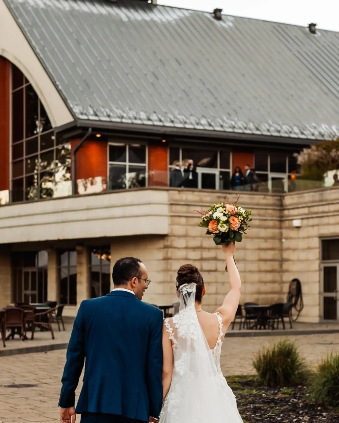 Un couple de mariés marche devant la salle de réception Plaza Rive Sud, une catégorie salle de réception renommée pour les événements de mariage. La mariée, en robe blanche élégante avec un voile, lève son bouquet de fleurs colorées vers le ciel. Le marié, en costume bleu, marche à ses côtés. Le bâtiment en arrière-plan présente une architecture moderne avec de grandes fenêtres et un toit métallique, offrant un cadre idéal pour célébrer des occasions spéciales comme un mariage.