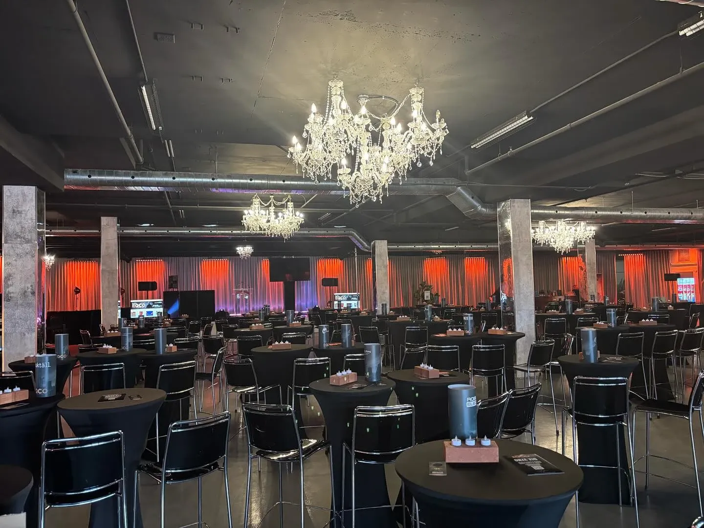 La salle de réception Plaza Rive Sud est magnifiquement aménagée pour un événement de mariage. La pièce spacieuse est ornée de lustres élégants suspendus au plafond, qui apportent une touche de sophistication. De nombreuses tables hautes avec des sièges noirs sont harmonieusement disposées pour accueillir les invités. Les murs sont illuminés de lumières rouges, créant une atmosphère chaleureuse et romantique, idéale pour une célébration de mariage.