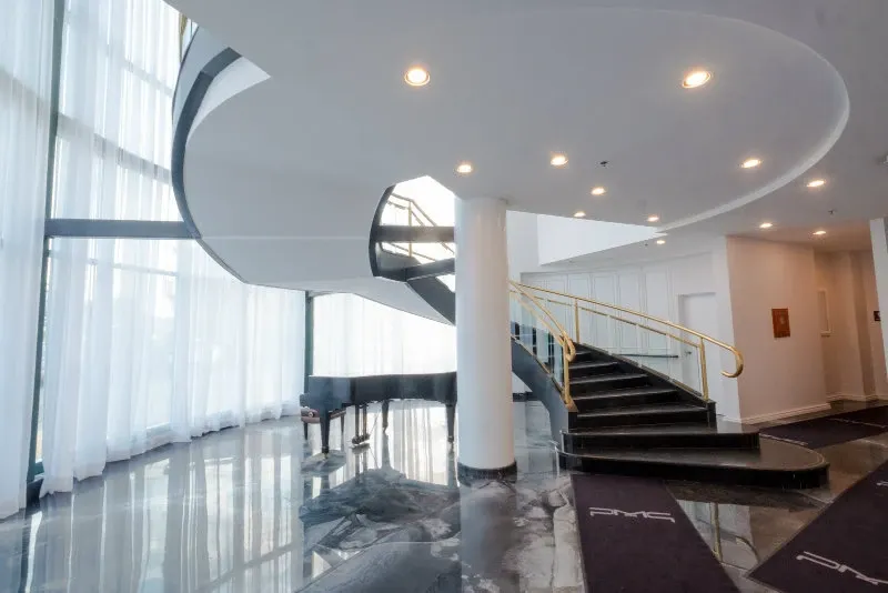 The image showcases the elegant interior of Plaza Vogue, a premier wedding venue under the category of Wedding Venues, featured on Mariage Elegant, Canada. It captures a sweeping, modern spiral staircase with a sleek black handrail, accompanied by a polished black grand piano placed near the base. The setting is illuminated by soft, natural light streaming through large, sheer curtain-covered windows, highlighting the glossy marble-like floor. This sophisticated decor reflects an ideal location for hosting luxurious wedding events.