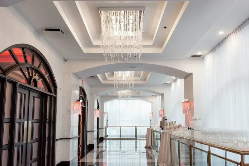 This image showcases a stylish interior view of the wedding venue Plaza Vogue, under the category of Wedding Venues, on Mariage Elegant, Canada. The venue features an elegant hallway with high, arched ceilings and luxurious chandeliers adorned with sparkling crystals that cast a soft glow. The walls are painted in a soothing white and are complemented by dramatic arched windows with dark wood frames. The sleek, polished flooring enhances the sophisticated ambiance. Tables along the side are draped in beige cloths, adding a touch of subtle elegance. This venue exudes a refined and modern aesthetic, ideal for a memorable wedding celebration.