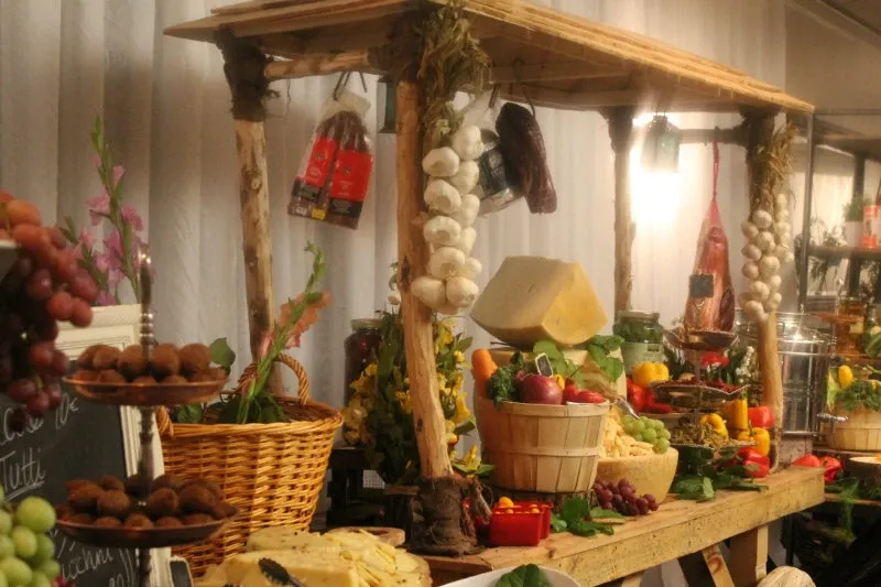 The image showcases an elegantly decorated display at Plaza Vogue, a featured vendor in the Wedding Venues category on Mariage Elegant, Canada. The setting includes a rustic wooden stall adorned with hanging garlic and cured meats, highlighting a rich selection of colorful fruits and vegetables in baskets, alongside a variety of cheese wheels. This visually appealing arrangement creates an inviting and sophisticated atmosphere ideal for a wedding reception, emphasizing the venue's commitment to quality and presentation.