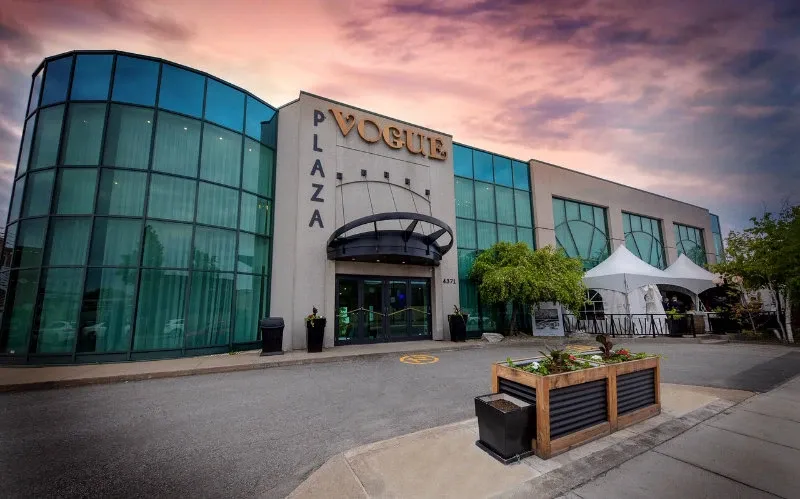 The image shows the exterior of Plaza Vogue, a prominent wedding venue in the Mariage Elegant vendor listing in Canada. The building features a modern design with large, curved glass windows and a stylish entrance canopy. A sign displaying 'VOGUE PLAZA' is prominently positioned above the entrance. The venue is surrounded by a well-maintained area with planters and a small patio, visible on the right. The sky is painted with hues of pink and purple, suggesting a serene and picturesque setting, ideal for weddings.