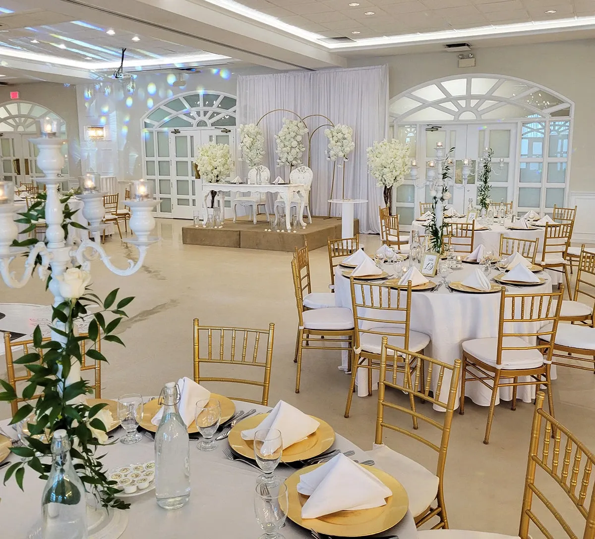 This image showcases the elegant wedding venue Plaza Vogue, categorized under Wedding Venues for Mariage Elegant in Canada. The venue features a beautifully decorated reception hall with round tables draped in white tablecloths, adorned with gold plates and neatly folded white napkins. Each table is surrounded by gold Chiavari chairs, enhancing the luxurious ambiance. The room is elegantly lit, with decorative chandeliers and soft, ambient lighting. In the background, a raised stage is set up with an ornate white backdrop and a floral arrangement, offering a perfect spot for wedding ceremonies or speeches. The overall setting exudes sophistication and charm, ideal for hosting a memorable wedding celebration.