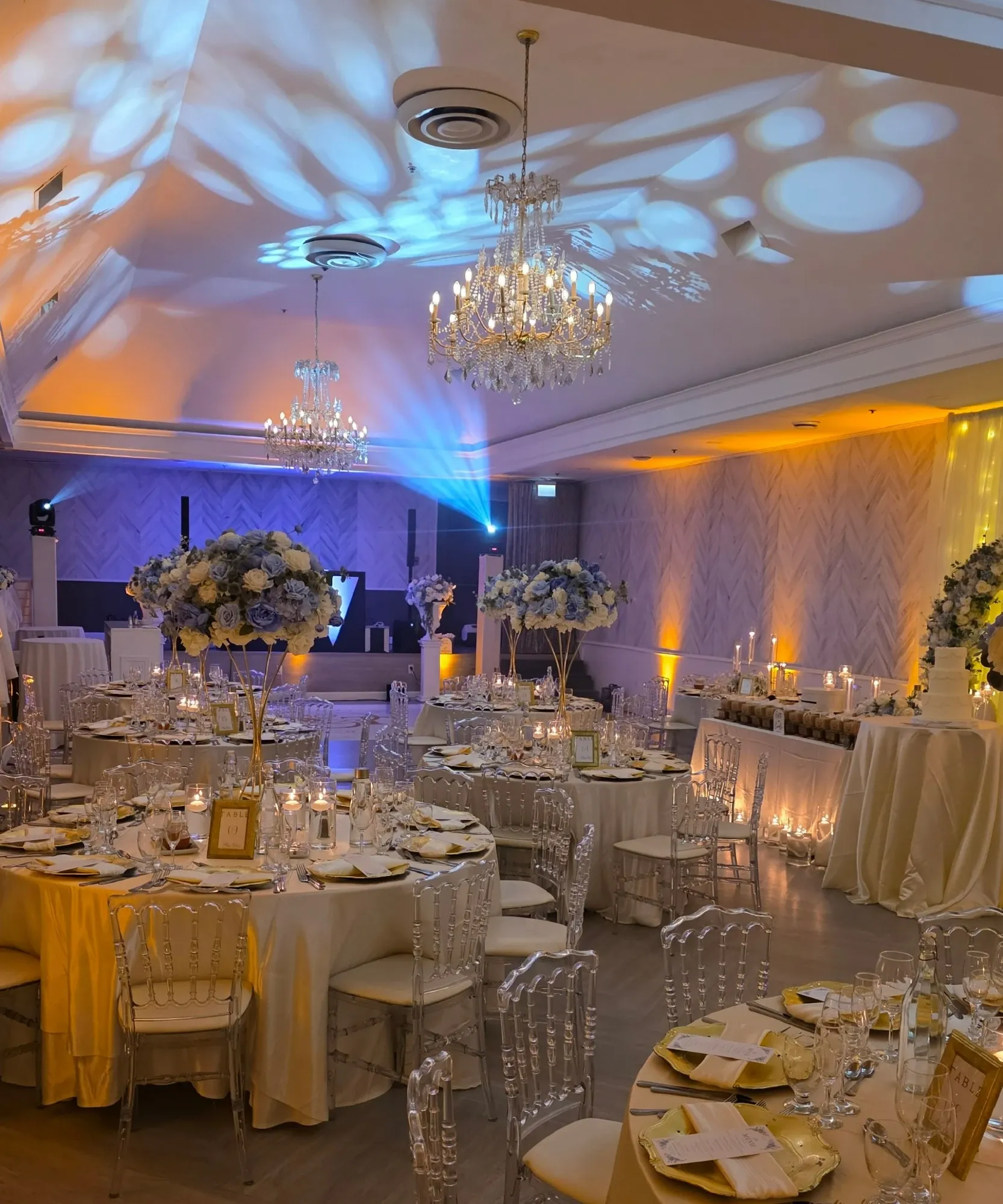 The image showcases the elegant wedding venue, Plaza Volare, a featured vendor on Mariage Elegant, Canada, under the Wedding Venues category. The venue is beautifully decorated, with round tables draped in ivory linen and set with gold-trimmed plates and crystal glassware. Tall, ornate floral centerpieces filled with white and blue flowers adorn each table. The room is softly lit by intricate crystal chandeliers hanging from the ceiling, creating a romantic ambiance. Wall-mounted lights cast gentle patterns across the room, enhancing the atmosphere. A tastefully arranged backdrop for the head table, featuring cascading yellow lights, adds a touch of glamour to the setting.