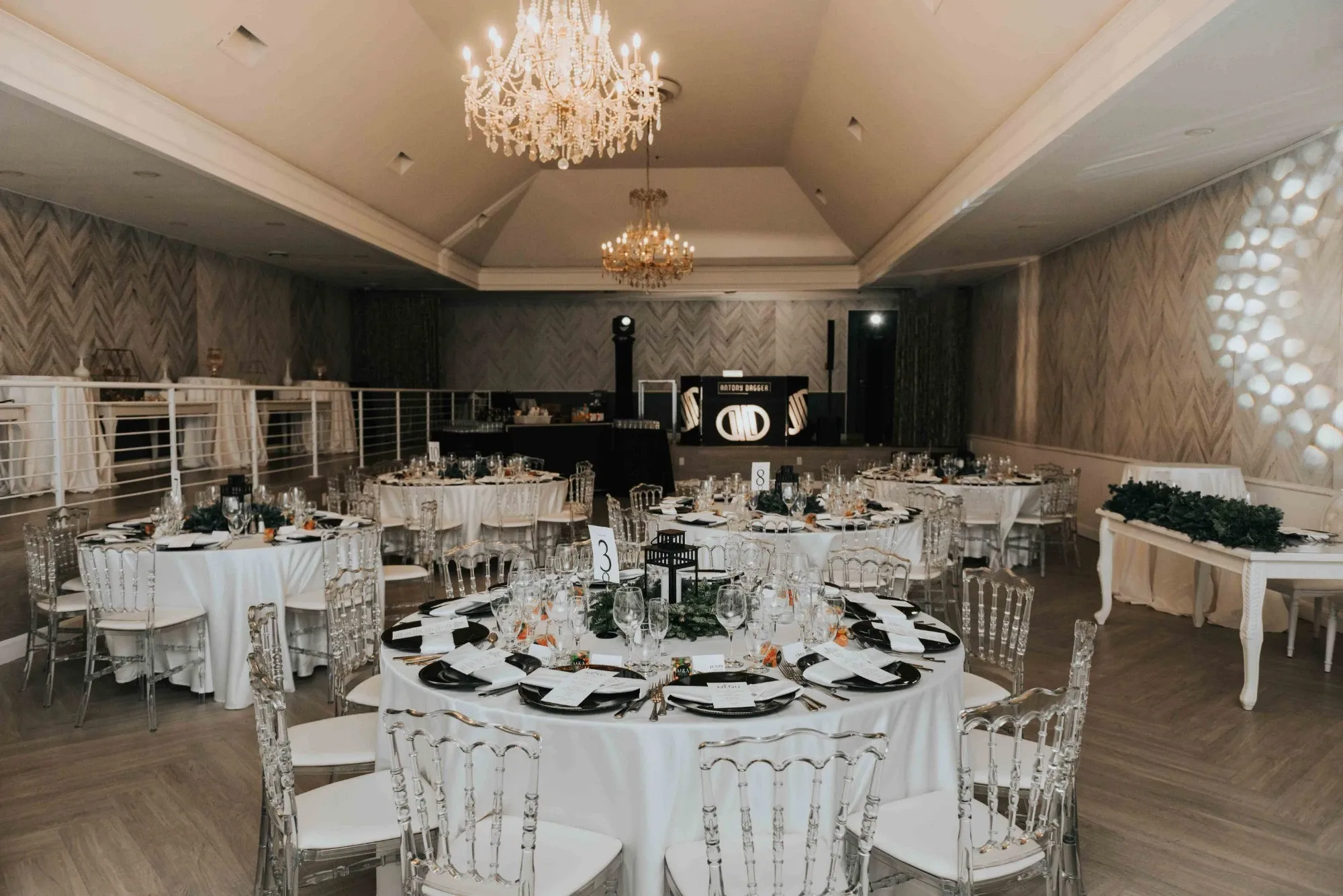 The image shows an elegantly decorated wedding venue at Plaza Volare, categorized under wedding venues. The spacious room features round tables adorned with white tablecloths and neatly arranged place settings, creating a sophisticated dining experience. Each table is accented with floral centerpieces and surrounded by translucent, decorative chairs. Above the scene, ornate chandeliers provide warm lighting, adding to the luxurious ambiance. The venue's ceiling has a vaulted design, enhancing the spacious feel. In the background, a stage area is set up, indicating space for entertainment or speeches during the wedding event.