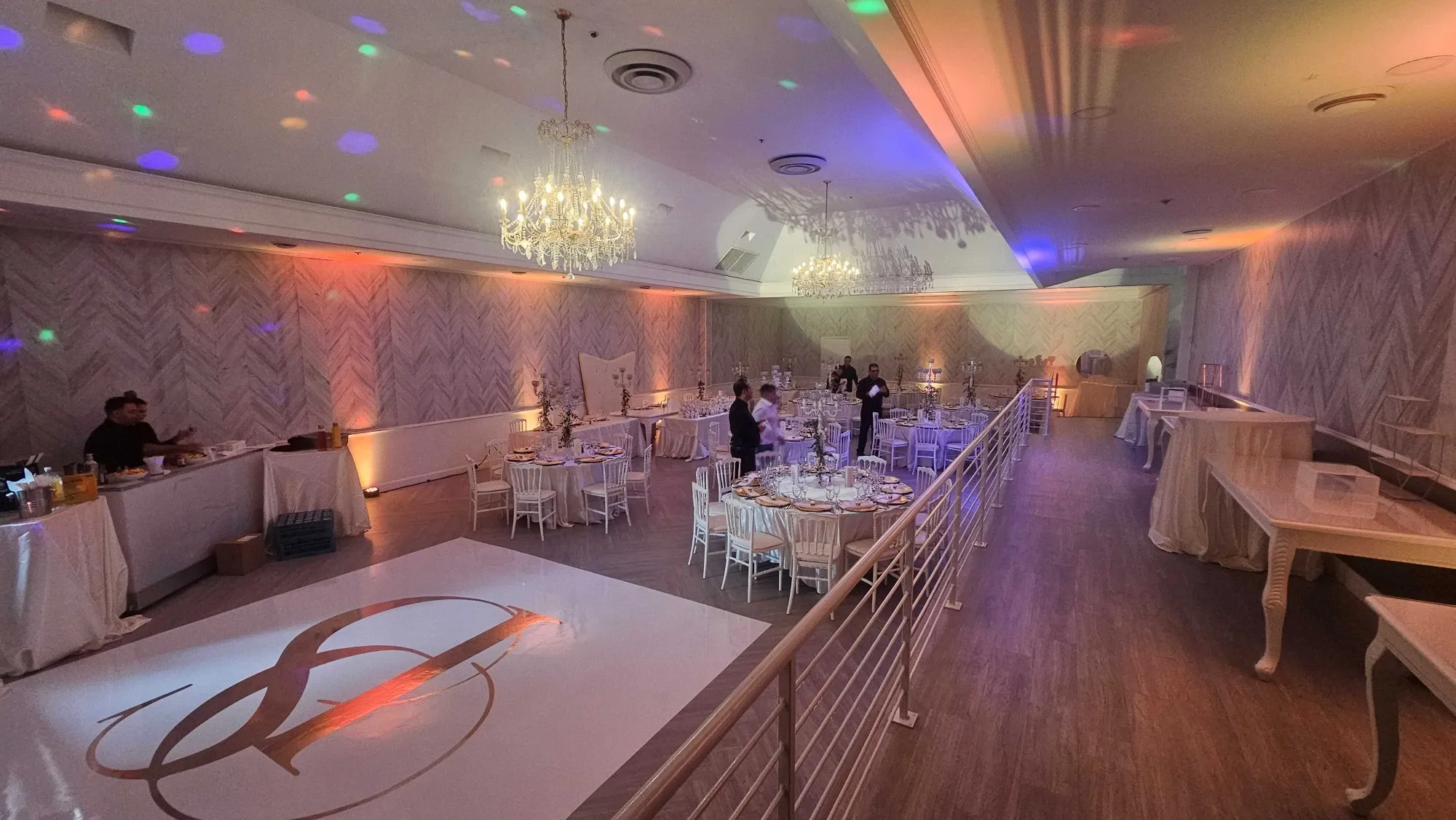 The image showcases Plaza Volare, a top-tier wedding venue featured on Mariage Elegant in Canada. The venue's interior is elegantly decorated, featuring crystal chandeliers that illuminate the spacious hall. Round tables are meticulously set with fine china and glassware, ready for a wedding celebration. The wooden floor boasts a prominent monogram, adding a personalized touch. The space is bathed in ambient lighting with subtle hues of orange and blue, creating a romantic atmosphere. Overall, the venue offers a sophisticated and enchanting setting suitable for hosting memorable wedding events.