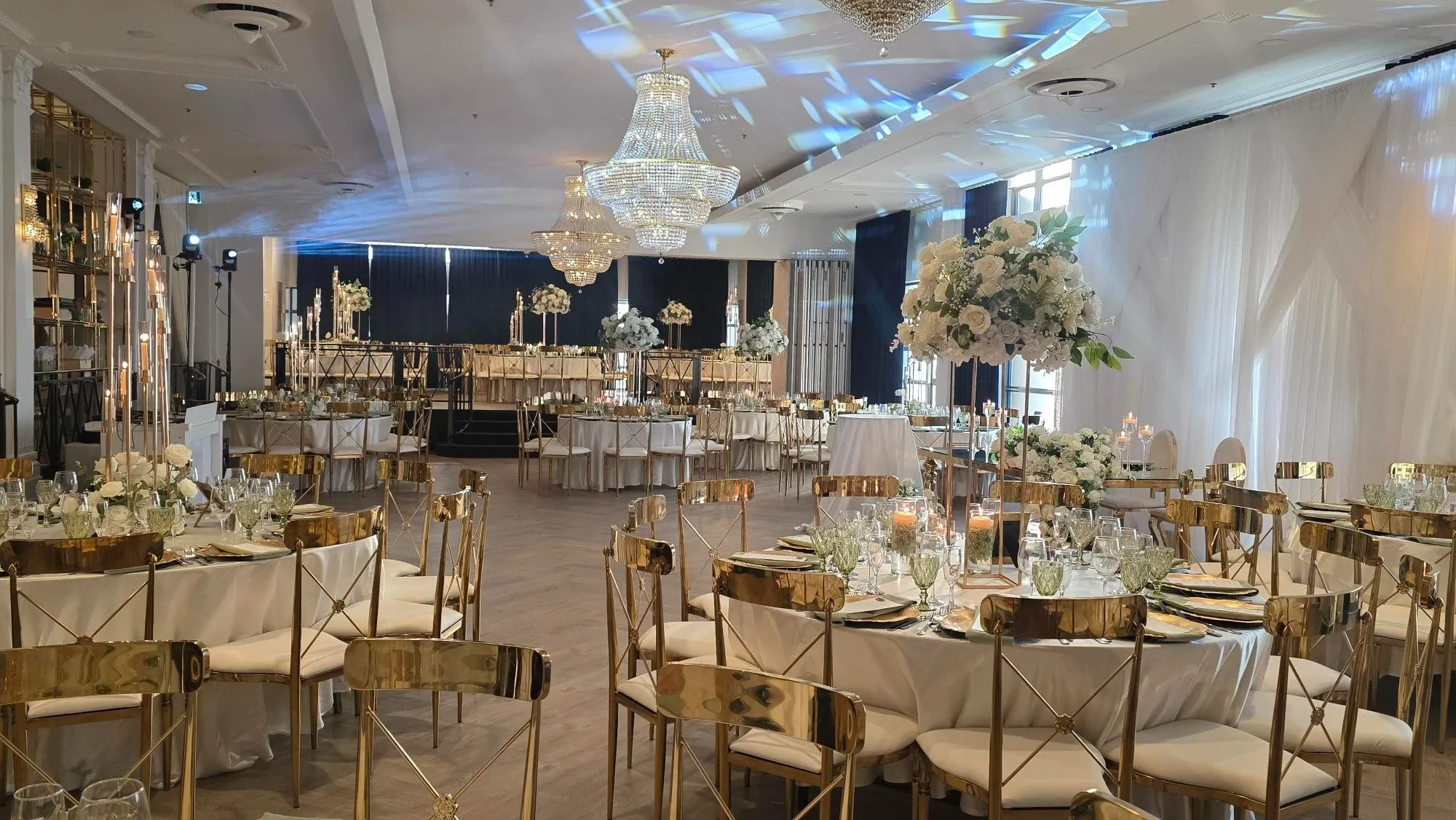 The image displays an elegantly decorated wedding venue at Plaza Volare, categorized under Wedding Venues for Mariage Elegant in Canada. The spacious room features round tables adorned with white tablecloths, surrounded by gold chairs. Each table is beautifully set with plates, glasses, and white floral centerpieces. The ceiling is highlighted by stunning crystal chandeliers, casting a warm glow over the setting. Large floral arrangements with white roses are prominently displayed, enhancing the luxurious atmosphere. In the background, sheer drapes and additional decorative lighting create a sophisticated ambiance, perfect for a memorable wedding celebration.