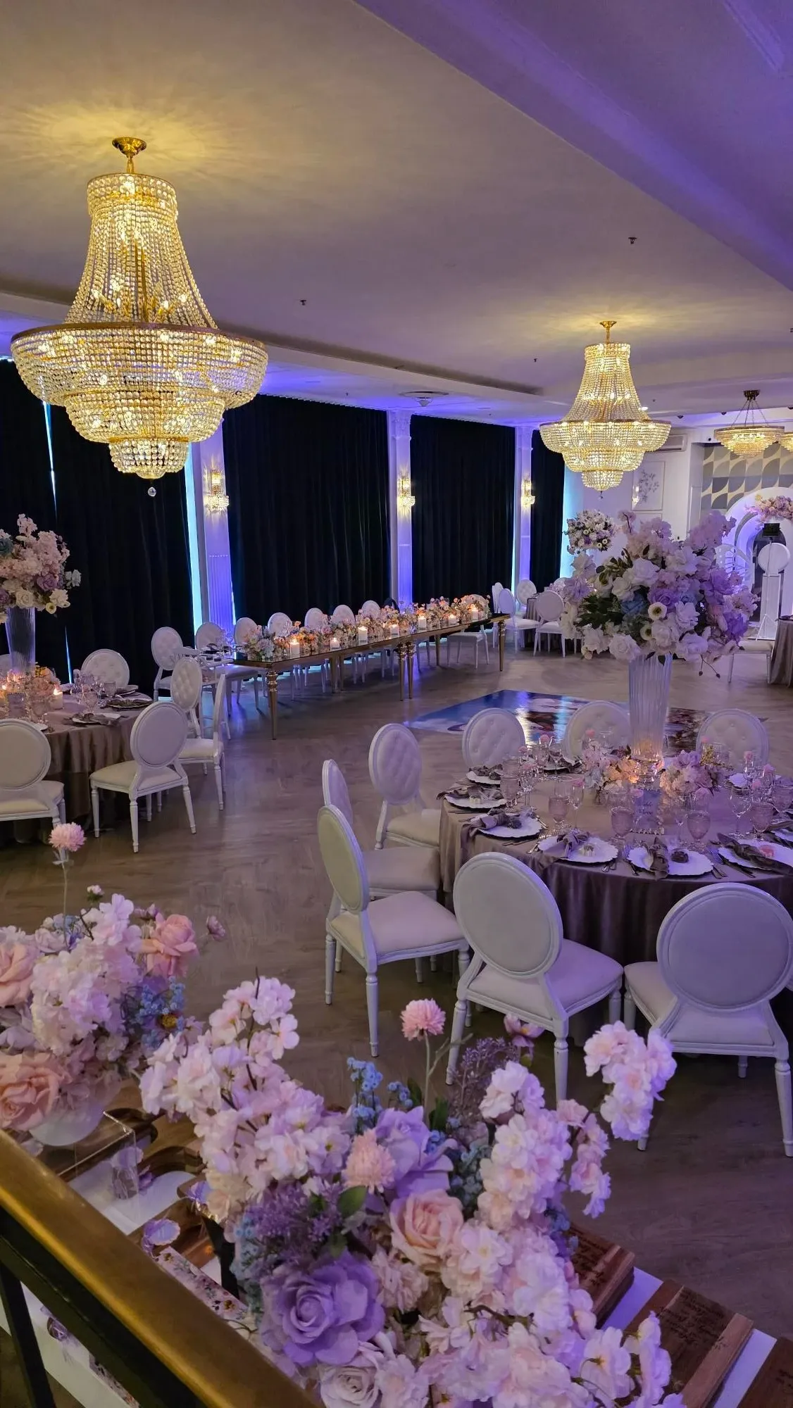 The image showcases a beautifully decorated wedding venue at Plaza Volare, categorized under Wedding Venues on Mariage Elegant, Canada. The elegant ballroom is adorned with large, sparkling chandeliers that illuminate the room with a soft, warm glow. Round tables are elegantly set with fine china and glassware, each surrounded by white cushioned chairs. The tables feature luxurious floral centerpieces with an abundance of pink and white flowers, adding a romantic ambiance. The walls are draped with dark curtains, providing an intimate atmosphere, and the floor is polished wood. This setting offers a sophisticated and enchanting space ideal for weddings.