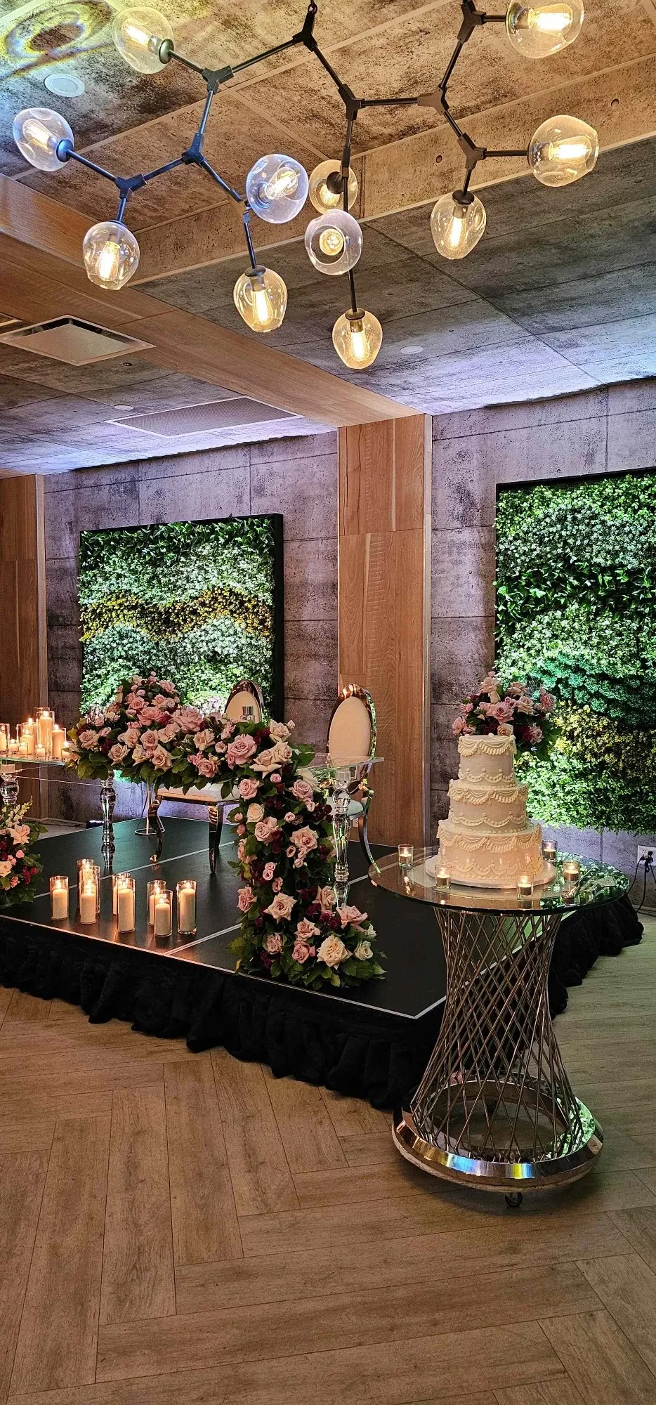 The image showcases an elegant wedding venue setup at Plaza Volare, categorized under wedding venues for Mariage Elegant in Canada. The scene features a beautifully decorated platform with two ornate chairs surrounded by lush floral arrangements in pink and white hues. A tastefully designed multi-tiered wedding cake rests on a stylish metallic stand nearby. Numerous small candlelights cast a warm glow, enhancing the romantic ambiance. The backdrop consists of green foliage panels, adding a natural touch to the sophisticated setting. Overhead, a modern chandelier with multiple glowing bulbs provides soft lighting, complementing the luxurious feel of the venue.