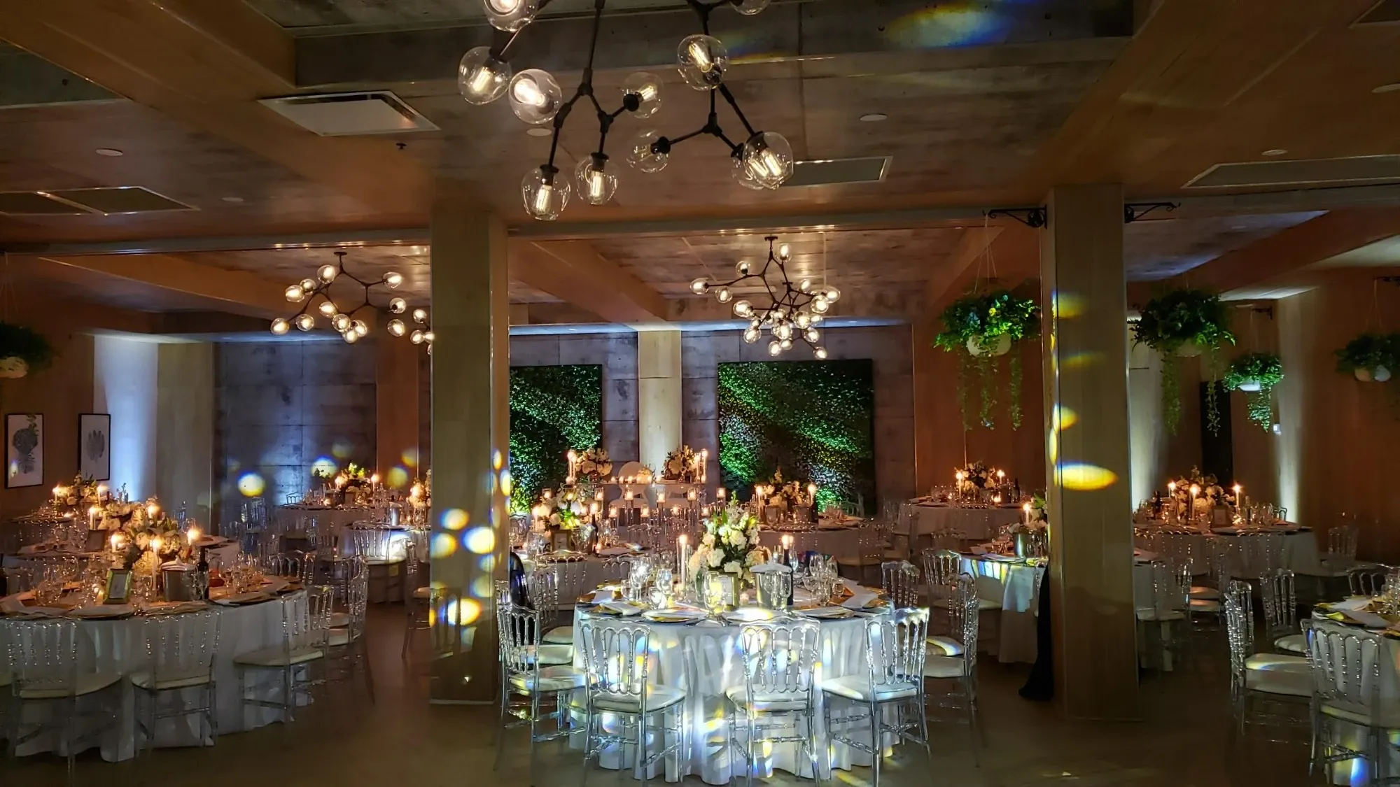 The image showcases an elegantly decorated wedding venue, Plaza Volare, part of Mariage Elegant's vendor listings in Canada. The spacious room is adorned with numerous round tables set with white linens and surrounded by clear, modern chairs. Each table features an intricate floral centerpiece with white and peach flowers, adding a touch of sophistication. Warm ambient lighting emanates from unique, modern chandeliers hanging from the ceiling, casting a romantic glow over the room. The backdrop includes lush greenery, contributing to the venue's inviting atmosphere, perfect for a memorable wedding celebration.
