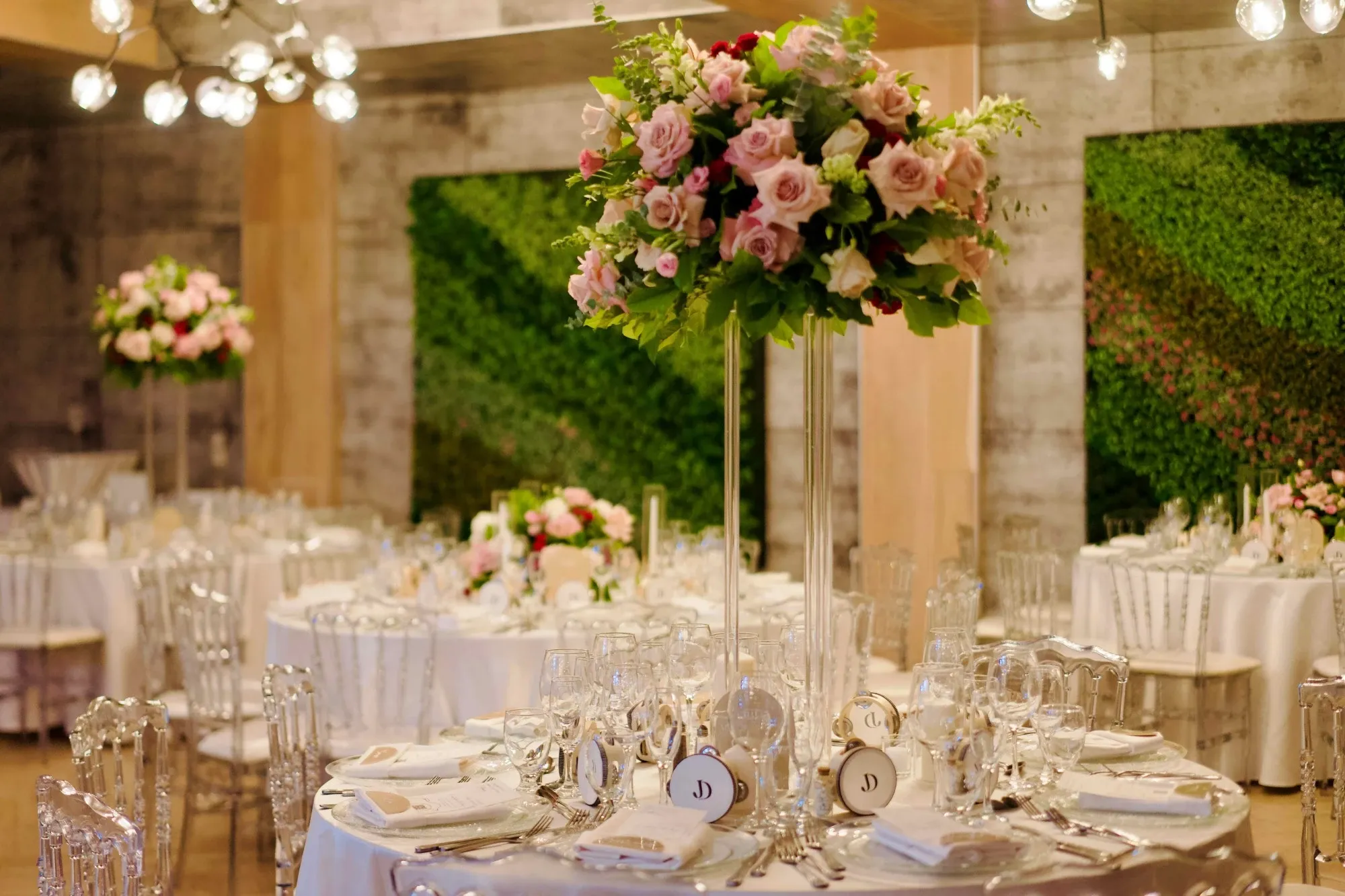 The image showcases an elegantly set wedding reception at Plaza Volare, a distinguished wedding venue in Canada. The tables are adorned with luxurious white tablecloths and are meticulously arranged with fine china, glassware, and silverware, creating an opulent dining experience. In the center, there is a tall, sophisticated floral arrangement featuring pink roses and lush greenery, adding a romantic touch to the setting. Soft lighting enhances the ambiance, reflecting off crystal chandeliers and the clear glasses, while a vibrant green wall in the background adds a hint of modern elegance to the venue.