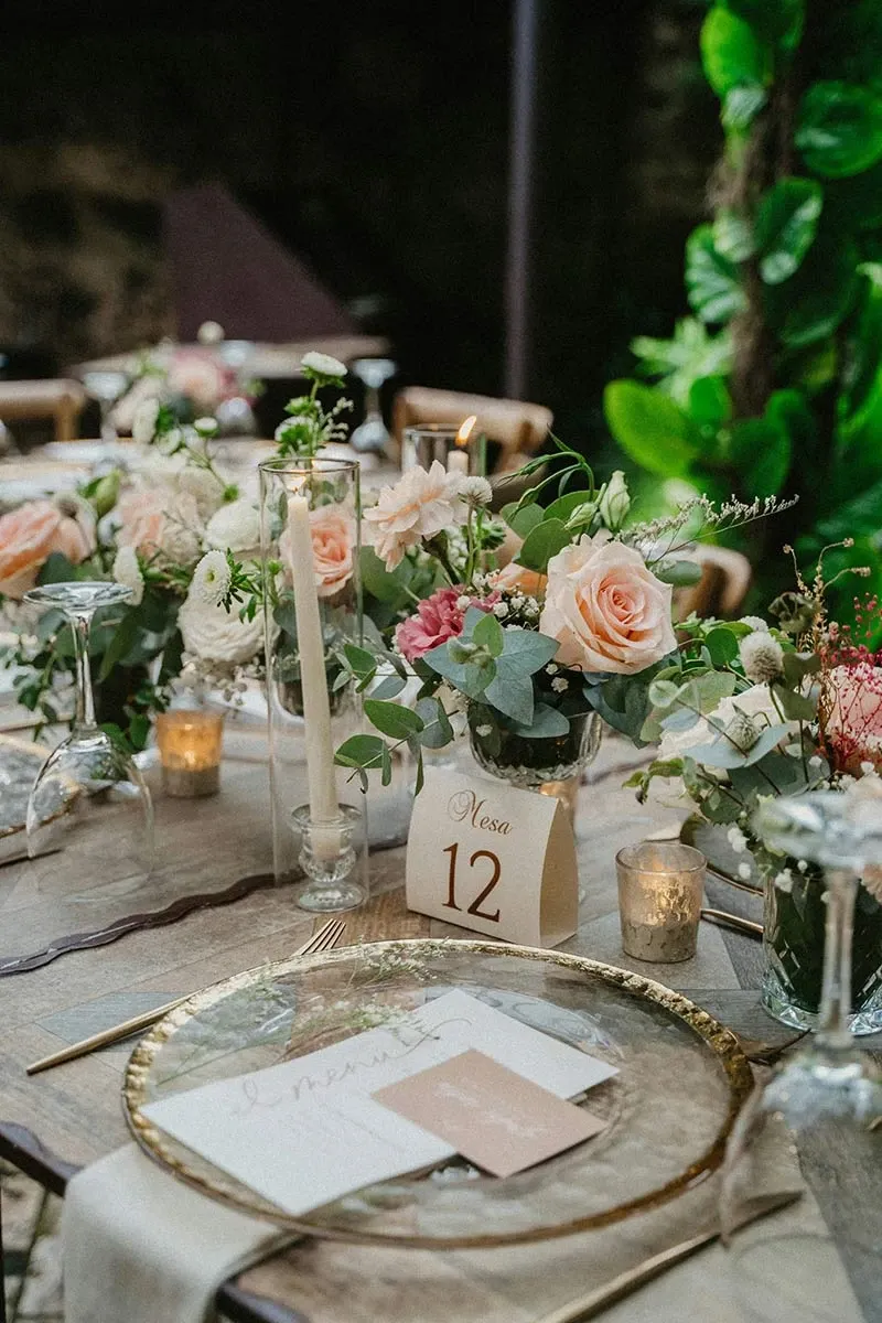 La photo montre une table de mariage élégamment décorée, idéale pour un événement chic. Au centre de la table, un bouquet de fleurs composé de roses pastel et de feuillage vert apporte une touche de fraîcheur. Les assiettes en verre avec des bords dorés sont disposées de manière sophistiquée, accompagnées d'une carte de place et d'un menu. Des bougies blanches, placées dans des chandeliers, ajoutent une ambiance romantique et chaleureuse. Un numéro de table 12 est visible, indiquant une organisation soignée pour le placement des invités. Dans l'arrière-plan, une végétation luxuriante renforce l’atmosphère naturelle et accueillante de la réception.