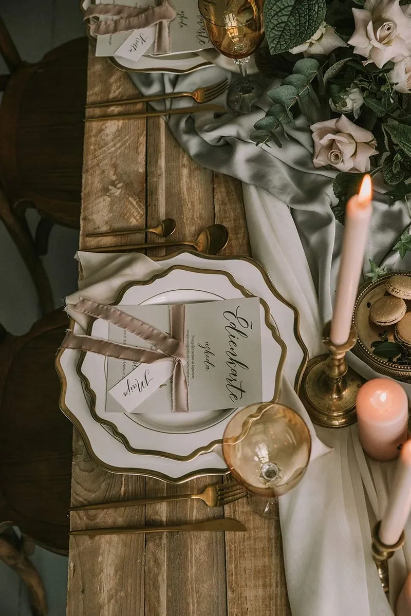 Une élégante table de banquet de mariage est dressée sur une table en bois rustique. La vaisselle présente des assiettes blanches à bord doré, avec des serviettes délicatement pliées et ornées de menus personnalisés. Des verres dorés ajoutent une touche glamour. Au centre, un chemin de table en tissu gris soyeux est décoré de fleurs blanches et de feuillage vert, complété par des bougies roses et blanches dans des chandeliers dorés. L'ambiance reflète un mariage raffiné et chic, idéal pour un mariage romantique au Canada.