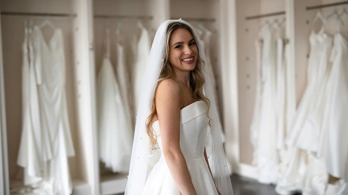 Une mariée souriante se tient dans une boutique de robes de mariée, entourée de plusieurs robes blanches élégantes suspendues en arrière-plan. Elle porte une robe de mariée moderne avec un voile délicat, reflétant une atmosphère de joie et d'anticipation. Cette image est idéale pour illustrer les services de planification de mariage proposés par Mariage Elegant, mettant en avant une sélection raffinée de tenues nuptiales.