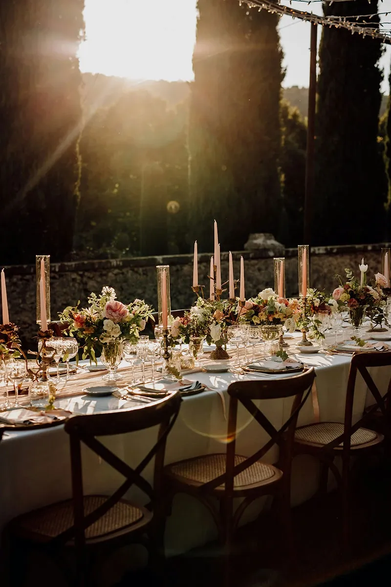 Une table de réception de mariage élégamment dressée est mise en lumière par un coucher de soleil pittoresque. La table est décorée de magnifiques arrangements floraux composés de roses, de dahlias et de verdure luxuriante, placés dans des vases en verre. De hauts chandeliers ornent la table, ajoutant une touche de sophistication. Les assiettes en porcelaine fine, les couverts en argent et les verres à vin complètent le décor raffiné. Des chaises en bois avec des coussins clairs entourent la table, créant une atmosphère chaleureuse et accueillante pour un mariage en plein air. La présence de grandes cyprès en arrière-plan ajoute un charme méditerranéen au cadre romantique.