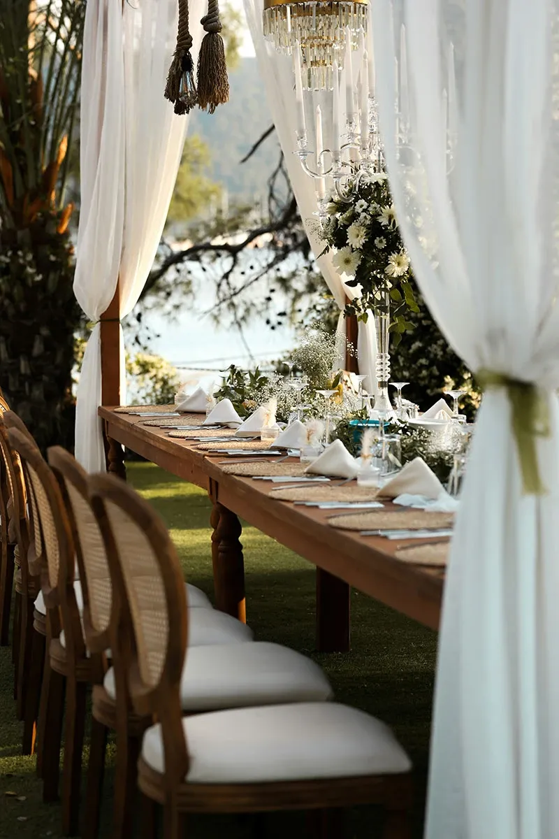 Une table de banquet élégamment dressée pour un mariage en plein air est visible dans l'image, ornée de nappes blanches et de serviettes pliées de manière raffinée. Les chaises classiques en bois avec des dossiers en osier complètent le décor sophistiqué. De magnifiques rideaux blancs flottent autour de la table, créant une ambiance intime et romantique. Un chandelier en cristal ajoute une touche de luxe, tandis que des arrangements floraux verdoyants rehaussent l'atmosphère naturelle de l'événement. L'arrière-plan montre un lac scintillant sous le soleil, accentuant le cadre idyllique de cette célébration nuptiale organisée par un planificateur de mariage professionnel au Canada.