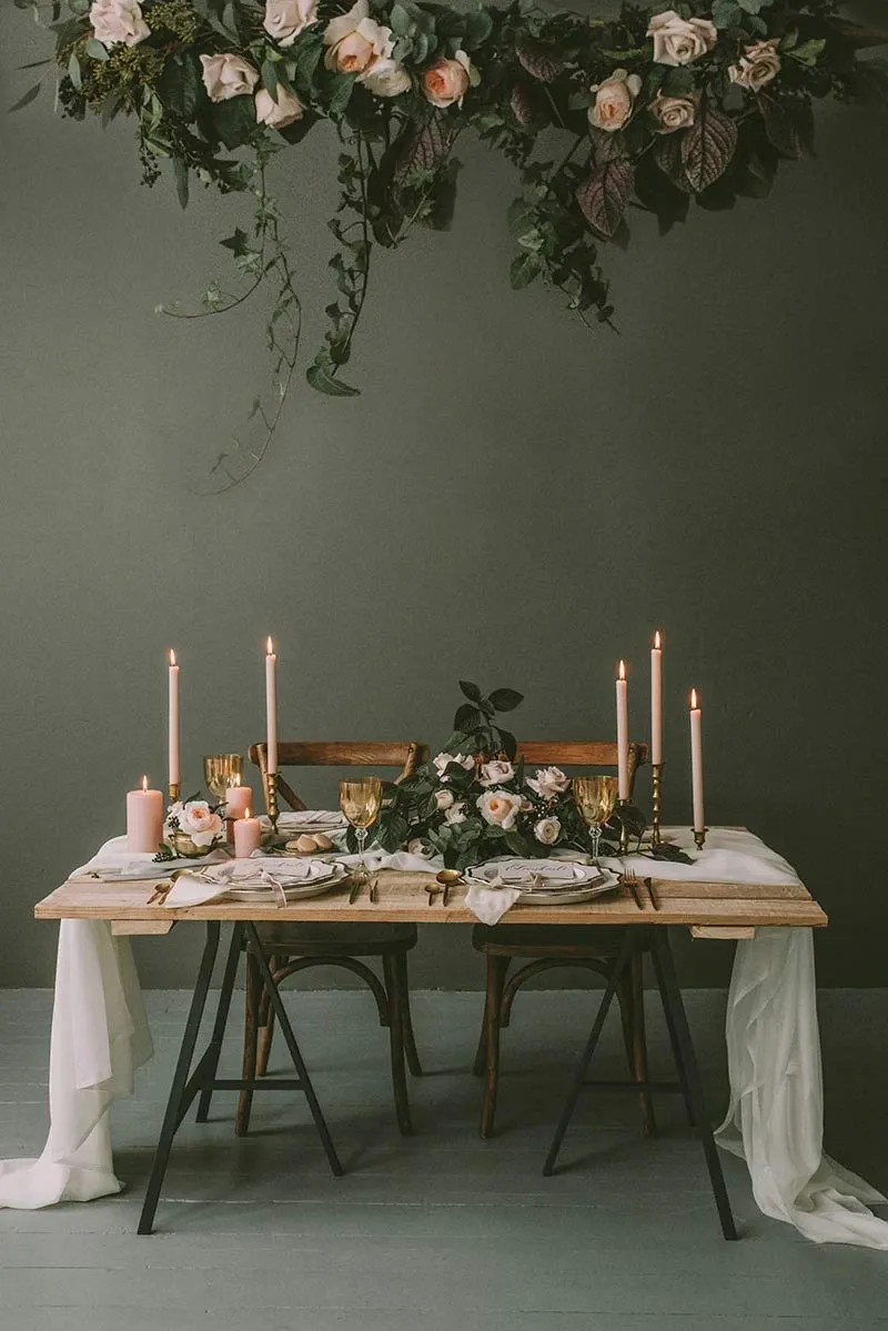 The image shows an elegantly set wedding table, featuring a rustic wooden table adorned with soft white drapes cascading over the sides. The centerpiece includes lush greenery and delicate pink roses, creating a romantic ambiance. Tall, slender candles in varying heights are symmetrically placed, with some lit to provide a warm, inviting glow. Gold-rimmed wine glasses and neatly arranged place settings add a touch of sophistication. Overhead, a floral arrangement of roses and leaves hangs, complementing the table decor. This setting exemplifies the meticulous attention to detail typical of expert wedding planners in Canada, perfect for a stylish and intimate wedding celebration.