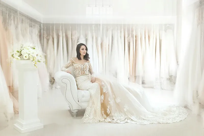 A bride sits gracefully on a plush white sofa in an elegantly decorated bridal boutique, surrounded by a backdrop of luxurious wedding dresses on display. She is wearing an exquisite lace wedding gown with intricate floral patterns and sheer sleeves, complemented by a long, flowing train. To her left, a tall white pedestal holds a beautiful floral arrangement with white roses and delicate greenery, adding to the sophisticated atmosphere. The setting exudes a sense of luxury and refinement, ideal for a premium wedding planning and vendor listing site like Mariage Elegant in Canada.