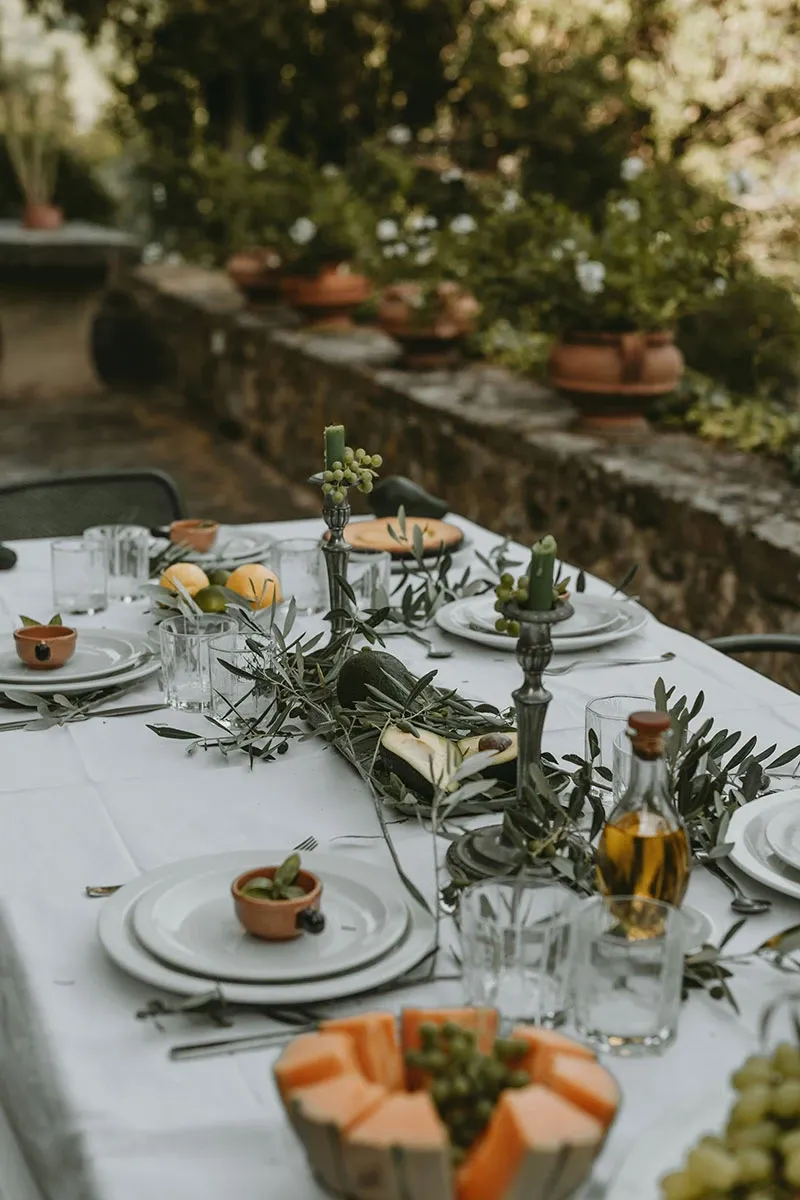 A beautifully set outdoor wedding table is adorned with elegant decor. The table features a pristine white tablecloth with neatly arranged white plates, silver cutlery, and clear glassware. Centerpieces include tall, dark candlesticks holding green candles and garlands of greenery with small fruits, adding a natural touch. A variety of fresh fruit, including slices of melon, is elegantly placed on the table for a refreshing touch. The background shows a rustic stone wall and lush greenery, creating an intimate and charming atmosphere, perfect for a wedding reception.