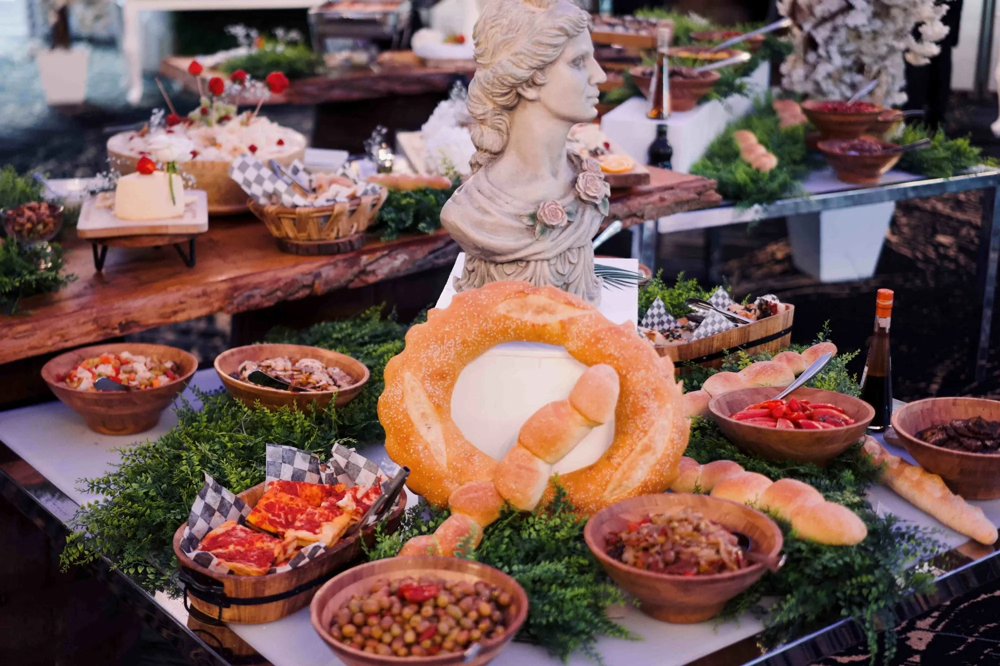 The image showcases an elegant buffet display at Plaza Volare, categorized under wedding venues for Mariage Elegant in Canada. The setup features a classic statue bust as the centerpiece, surrounded by a variety of appetizing dishes. There are wooden bowls filled with assorted olives, nuts, and roasted vegetables. A prominent bread arrangement encircles a cheese wheel, adding an artistic touch to the presentation. The table is adorned with lush greenery, enhancing the sophisticated atmosphere of this wedding venue.