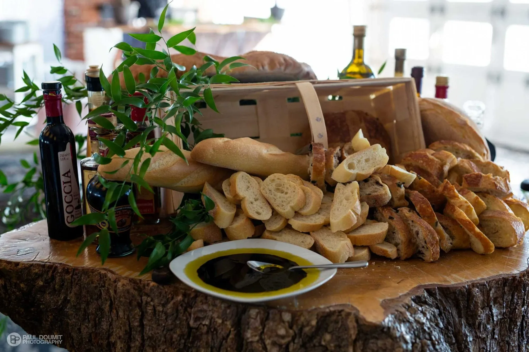 This image showcases an elegant array of assorted bread displayed on a rustic wooden table at Plaza Volare, a featured wedding venue on Mariage Elegant, Canada. The scene includes a wooden crate overflowing with various types of sliced baguettes and loaves, artfully arranged with green leafy branches for an organic touch. Surrounding the bread are bottles of olive oil and balsamic vinegar, adding a gourmet element suitable for wedding receptions. The setup exudes a warm and inviting atmosphere, ideal for a wedding celebration.