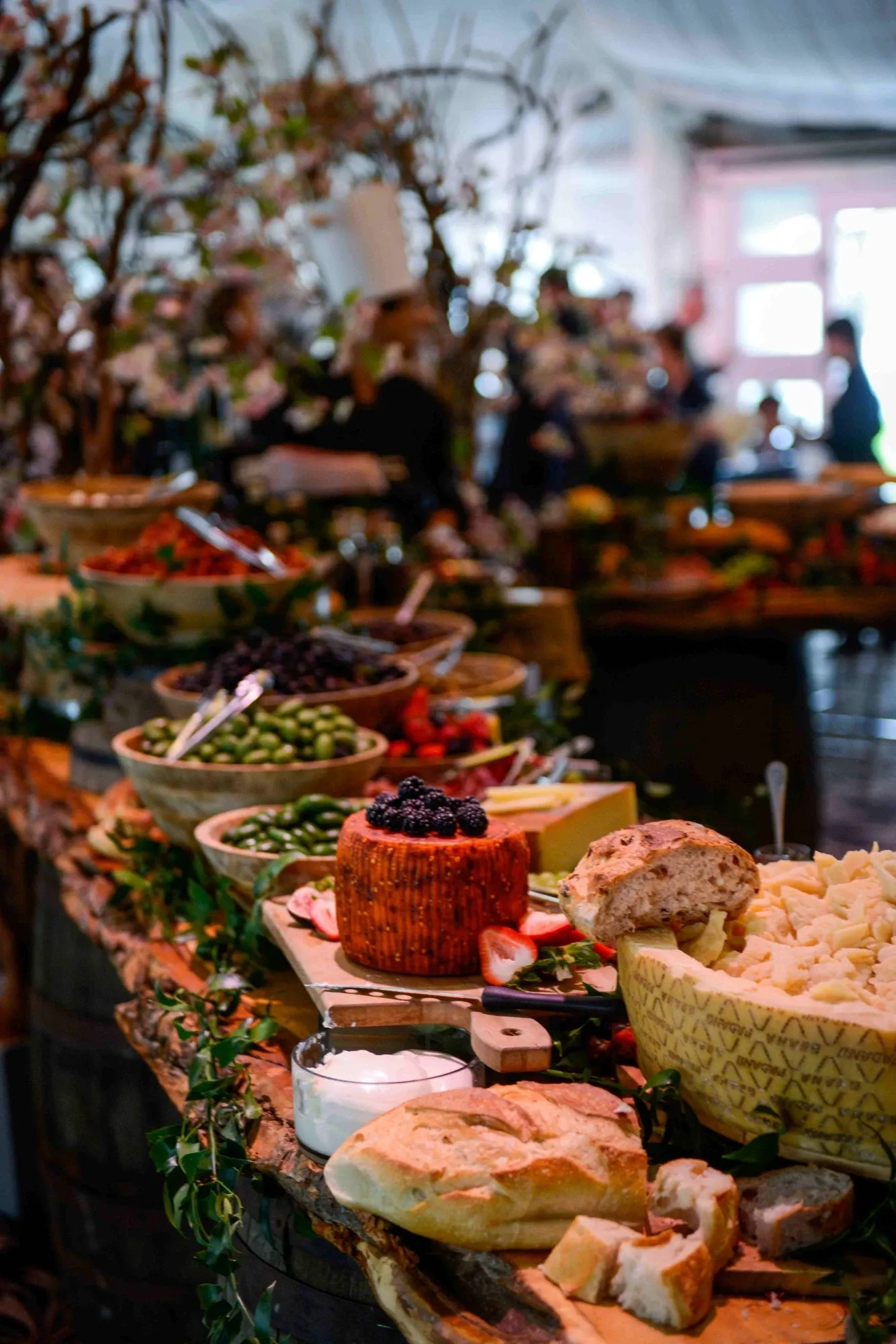 The image showcases an elegant wedding venue buffet setup at Plaza Volare, listed under the Wedding Venues category on Mariage Elegant, Canada. The beautifully arranged tables are adorned with an assortment of fresh, artisanal cheeses, olives, and breads. A prominent cheese wheel garnished with berries takes center stage, surrounded by leafy decorations, adding a rustic charm. The ambient lighting and soft-focus background suggest a warm, inviting atmosphere, ideal for wedding celebrations.