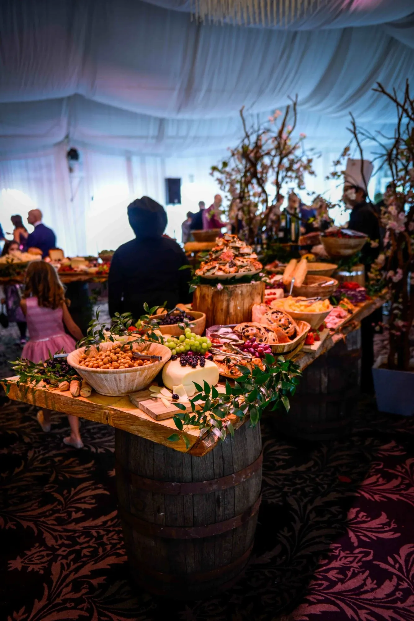 The image features an elegant wedding reception setup at Plaza Volare, a top wedding venue in Canada. A lavish display of gourmet snacks and appetizers is artfully arranged on rustic wooden barrels. The table boasts an assortment of cheeses, fresh grapes, assorted nuts, and beautifully arranged charcuterie. The venue is adorned with intricate floral arrangements and lush greenery, creating a charming and inviting atmosphere. Guests, including a child in formal attire, mingle in the background under soft, ambient lighting, encapsulating the perfect setting for a wedding celebration.