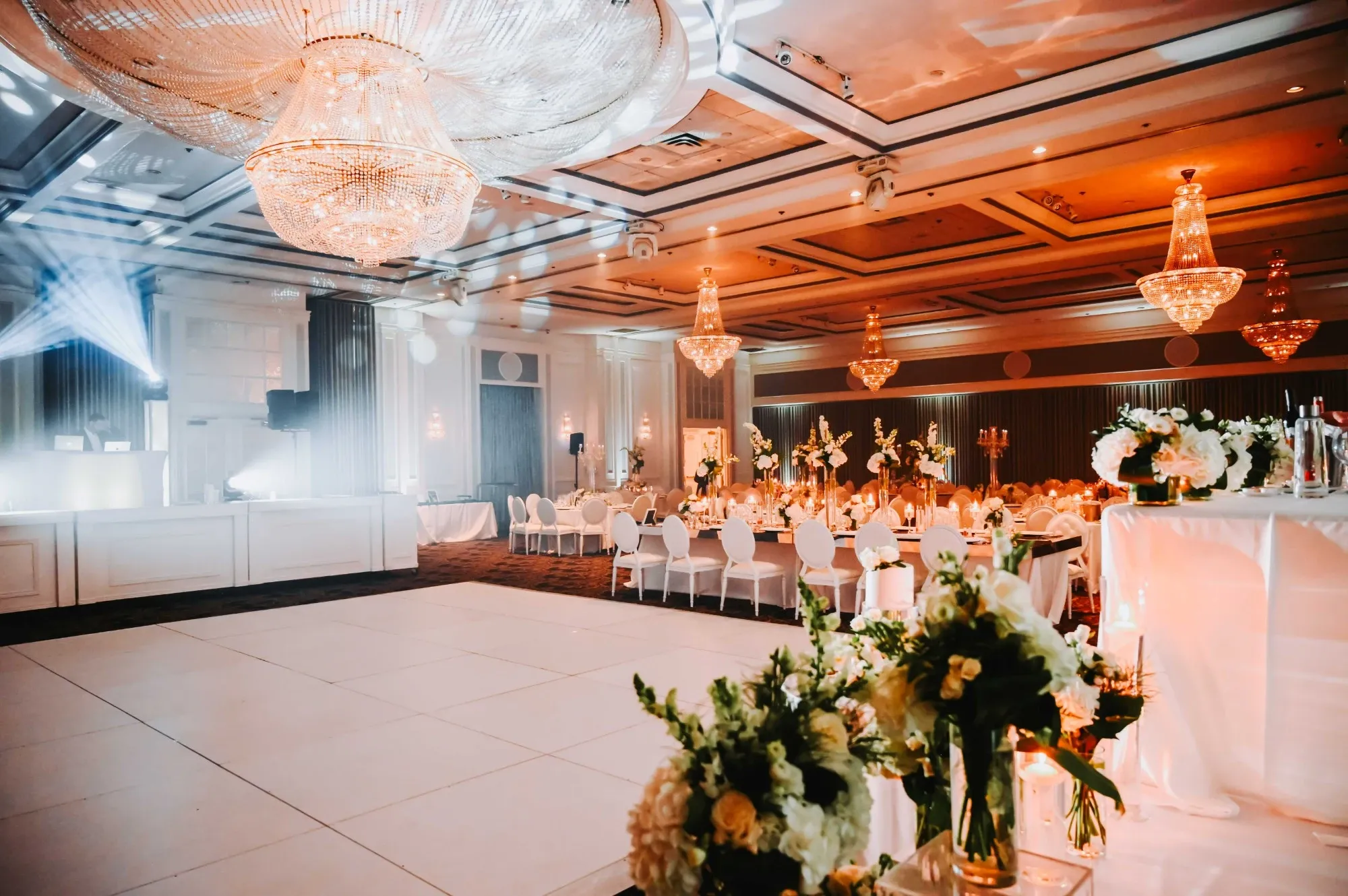 The image showcases an elegantly decorated wedding venue at Plaza Volare, categorized under Wedding Venues on the Mariage Elegant website. The spacious room features a large, glistening dance floor surrounded by beautifully arranged tables adorned with floral centerpieces. Luxurious chandeliers hang from the ceiling, casting a warm, ambient glow throughout the space. The setup also includes tastefully arranged lighting and floral arrangements. This venue highlights a sophisticated and inviting atmosphere, ideal for hosting weddings in Canada.