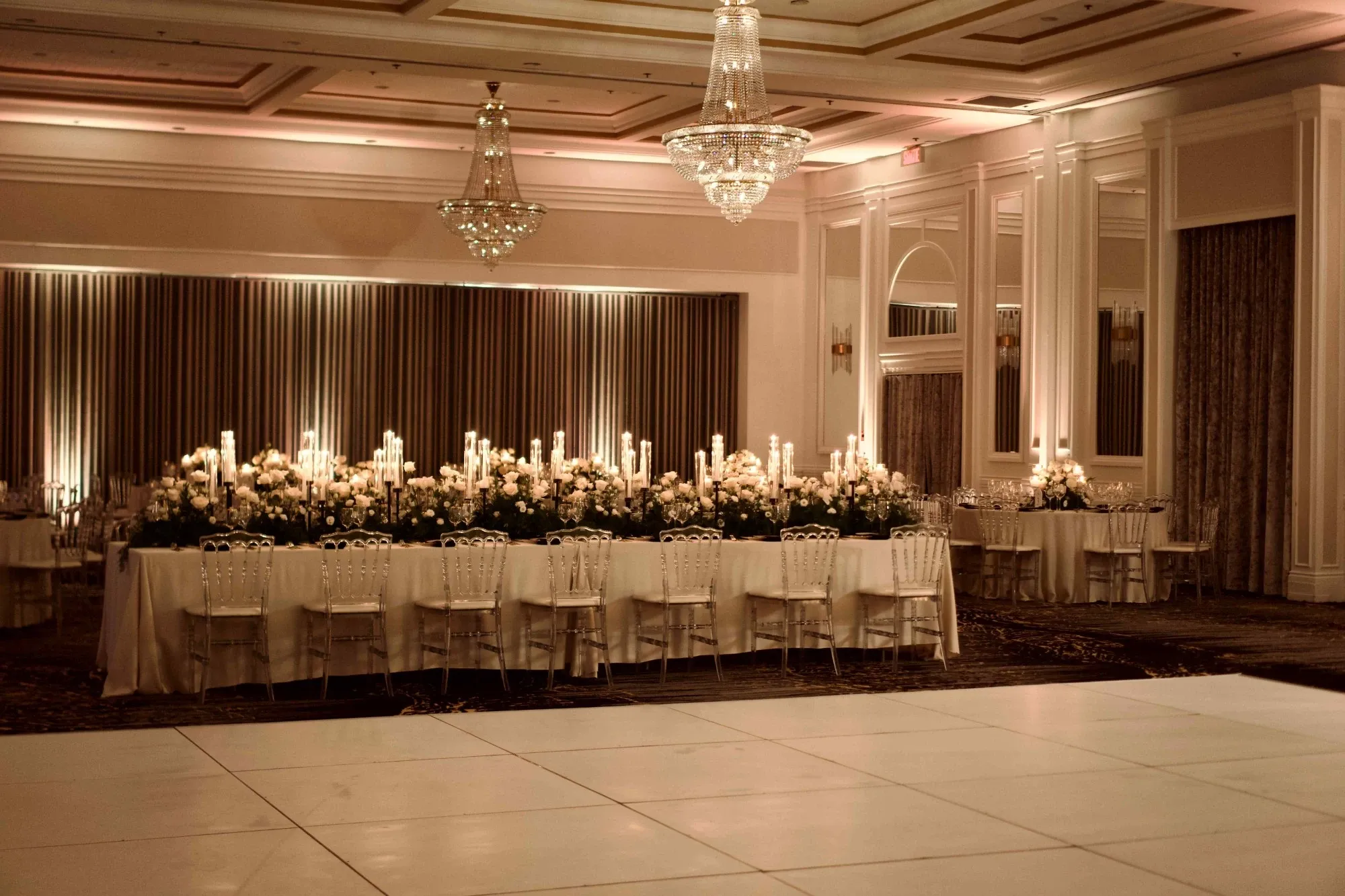 The image showcases an elegant wedding venue at Plaza Volare, categorized under wedding venues, located in Canada. The spacious banquet hall is beautifully set up with a long, rectangular table adorned with white tablecloths and elegantly arranged floral centerpieces. Numerous tall, lit candles add a romantic ambiance to the scene. The sophisticated decor includes grand chandeliers that hang from the ceiling, casting a warm glow over the venue. Tall drapes and mirrored walls enhance the opulence of the space, reflecting the light and creating an inviting atmosphere for wedding celebrations.