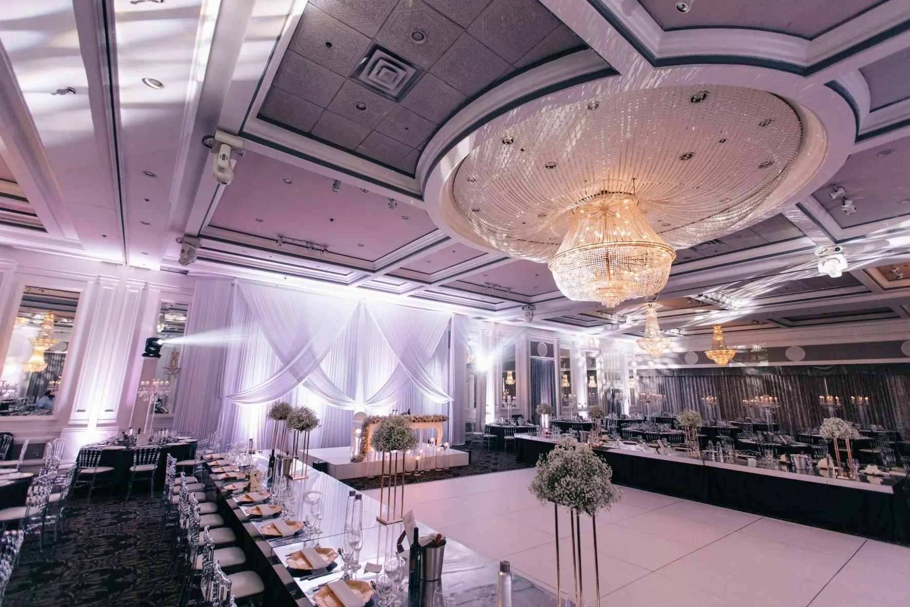 The image showcases the elegant interior of Plaza Volare, a premier wedding venue in the Mariage Elegant directory in Canada. The spacious hall is adorned with a magnificent central chandelier casting a warm glow over the room. The ceiling features intricate moldings and recessed lighting, enhancing the sophisticated ambiance. A large dance floor is centrally positioned, surrounded by beautifully arranged tables set with fine dinnerware and elegant floral centerpieces, ready to host a wedding reception. Luxurious draped fabric and soft lighting frame the head table, creating a romantic setting for the celebration.
