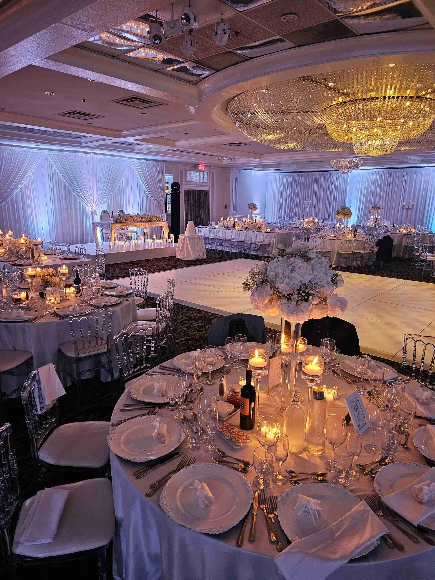 The image showcases an elegant wedding venue at Plaza Volare, categorized under Wedding Venues for Mariage Elegant in Canada. The venue features a beautifully decorated reception area, with round tables set for an event. Each table is adorned with pristine white tablecloths, neatly folded napkins, and sparkling glassware, creating a refined ambiance. In the center of the tables are tall, elegant floral centerpieces surrounded by glowing candles, adding a romantic touch. In the background, a lavishly illuminated dance floor is visible, under a stunning, large crystal chandelier that enhances the sophistication of the space. The soft, draped fabric along the walls and subtle, ambient lighting contribute to the luxurious atmosphere of this wedding venue.